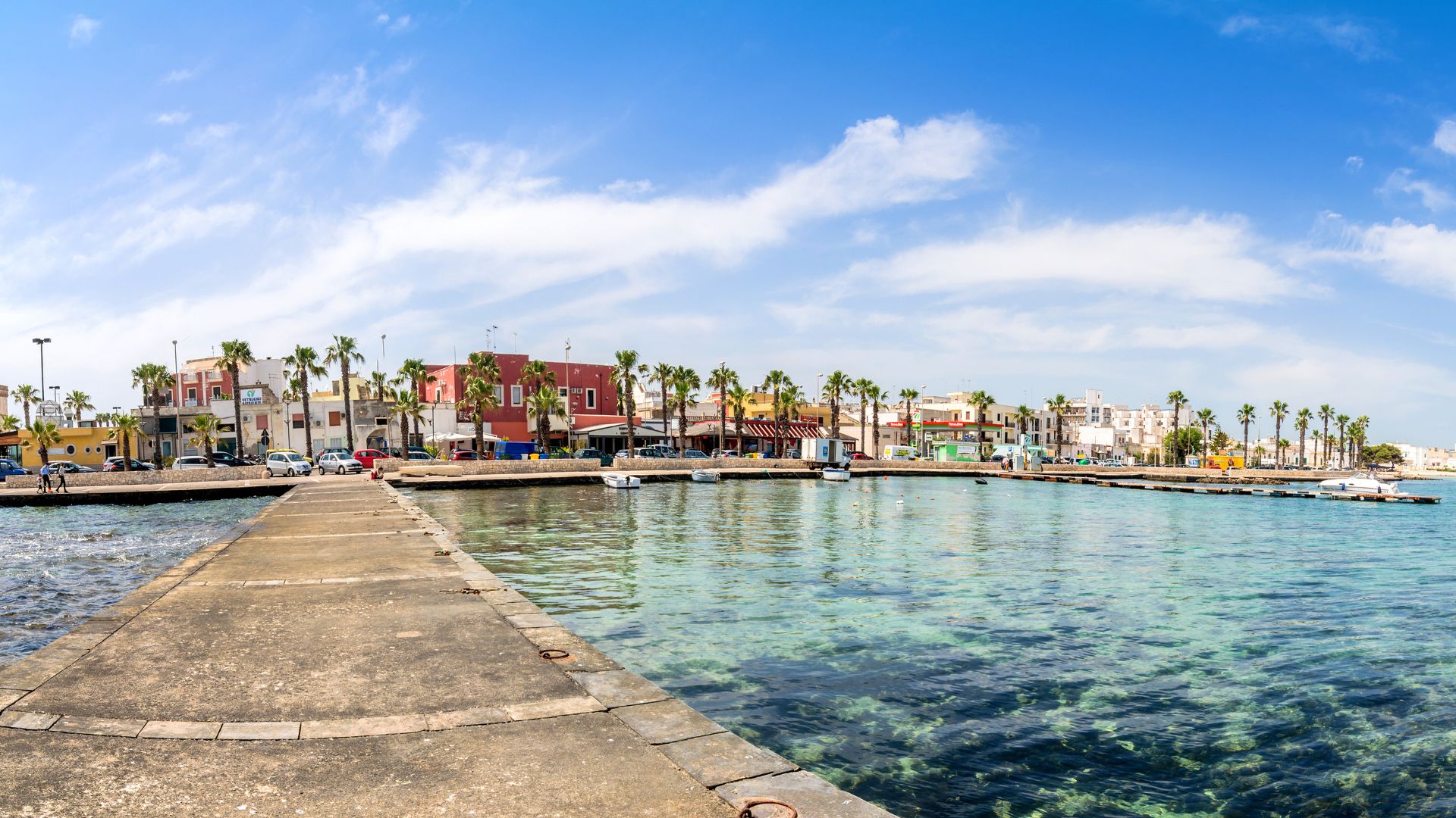The image shows the harbor area, with a concrete pier extending into the clear, shallow water, and the town's buildings lining the waterfront in Porto Cesareo, Italy