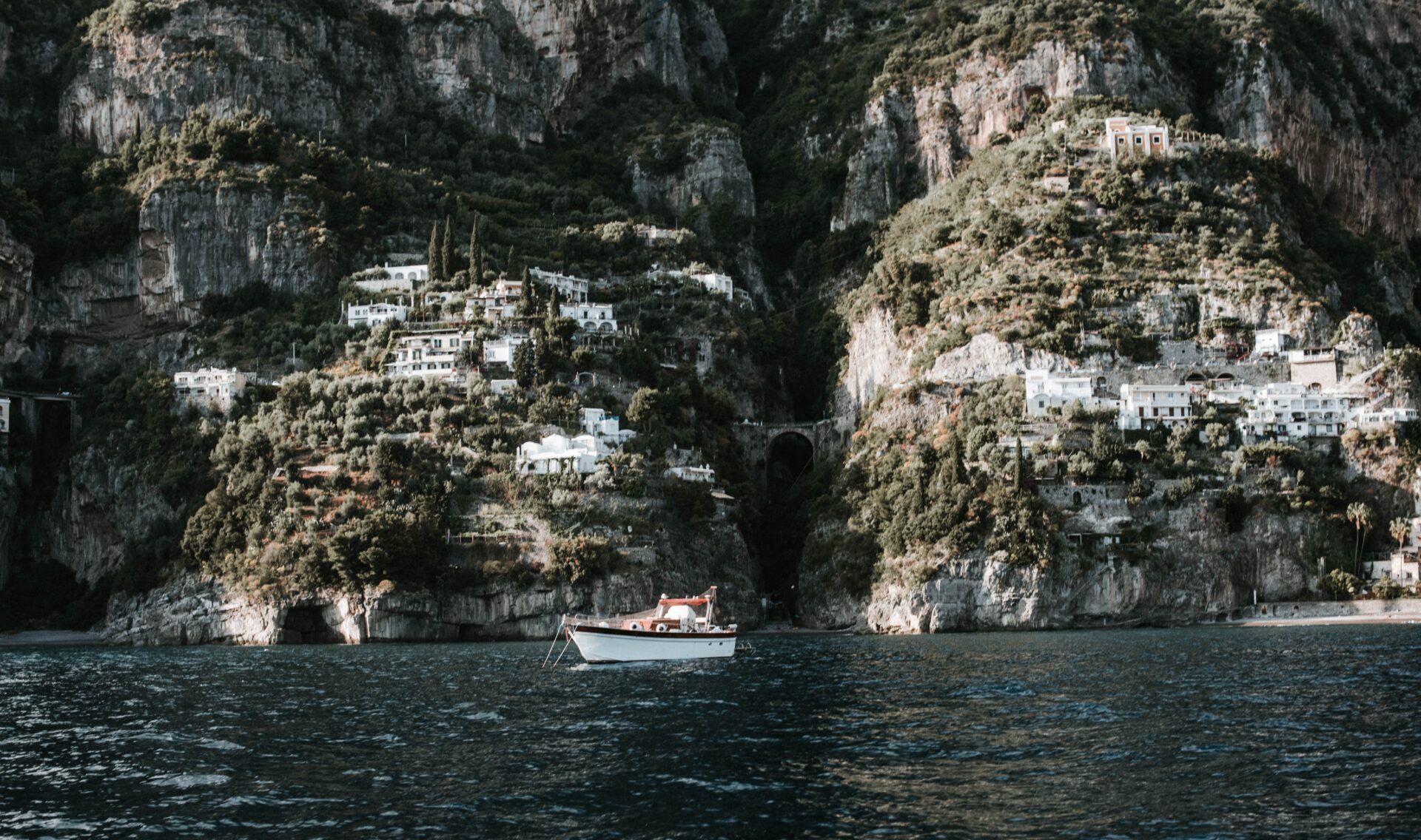 A scenic view of Praiano, Italy, with white houses nestled on green hillsides above the deep blue sea and a small boat floating in the water.