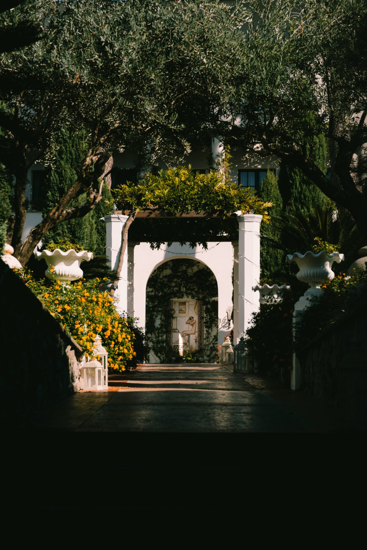 Shaded garden path leading to a white archway and wall art in Ravello, Italy, surrounded by trees and flowers.
