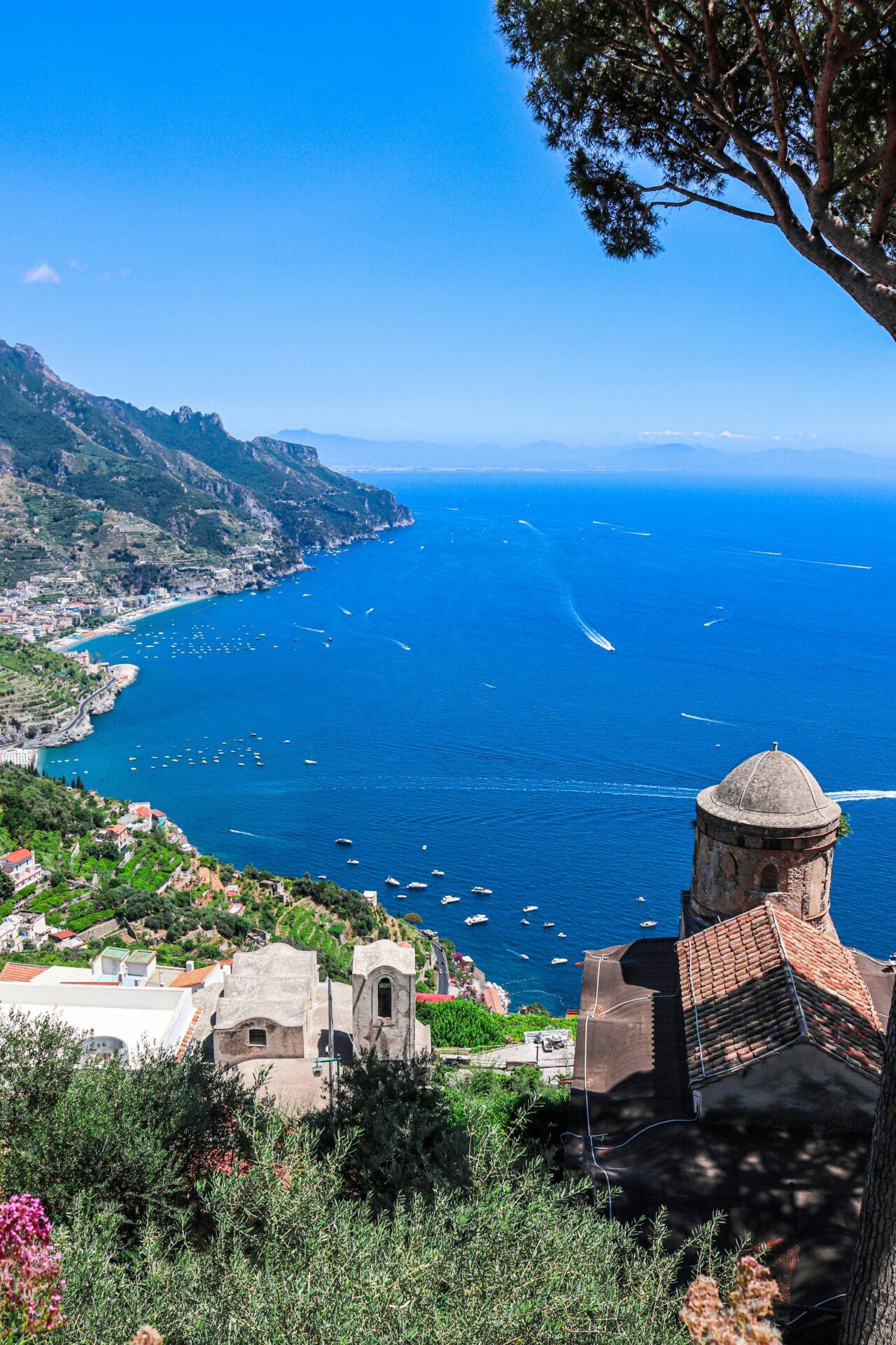 View of the Amalfi Coast from Ravello, Italy, with historic buildings, lush greenery, and boats scattered across the bright blue sea.