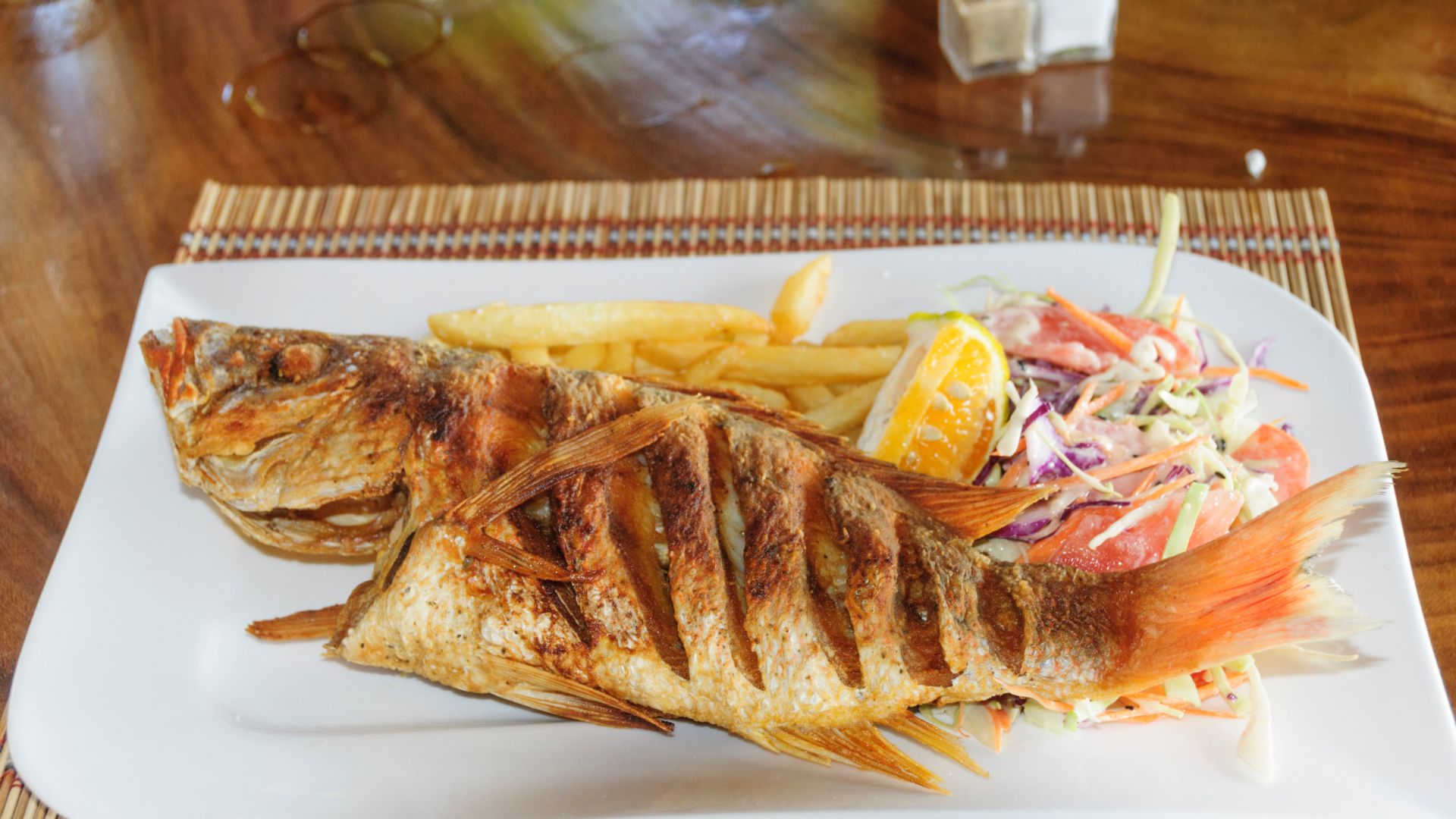 The image shows fried Red Snapper prepared whole, with slashes on its side. It is served with french fries, a lemon wedge, and a side salad.