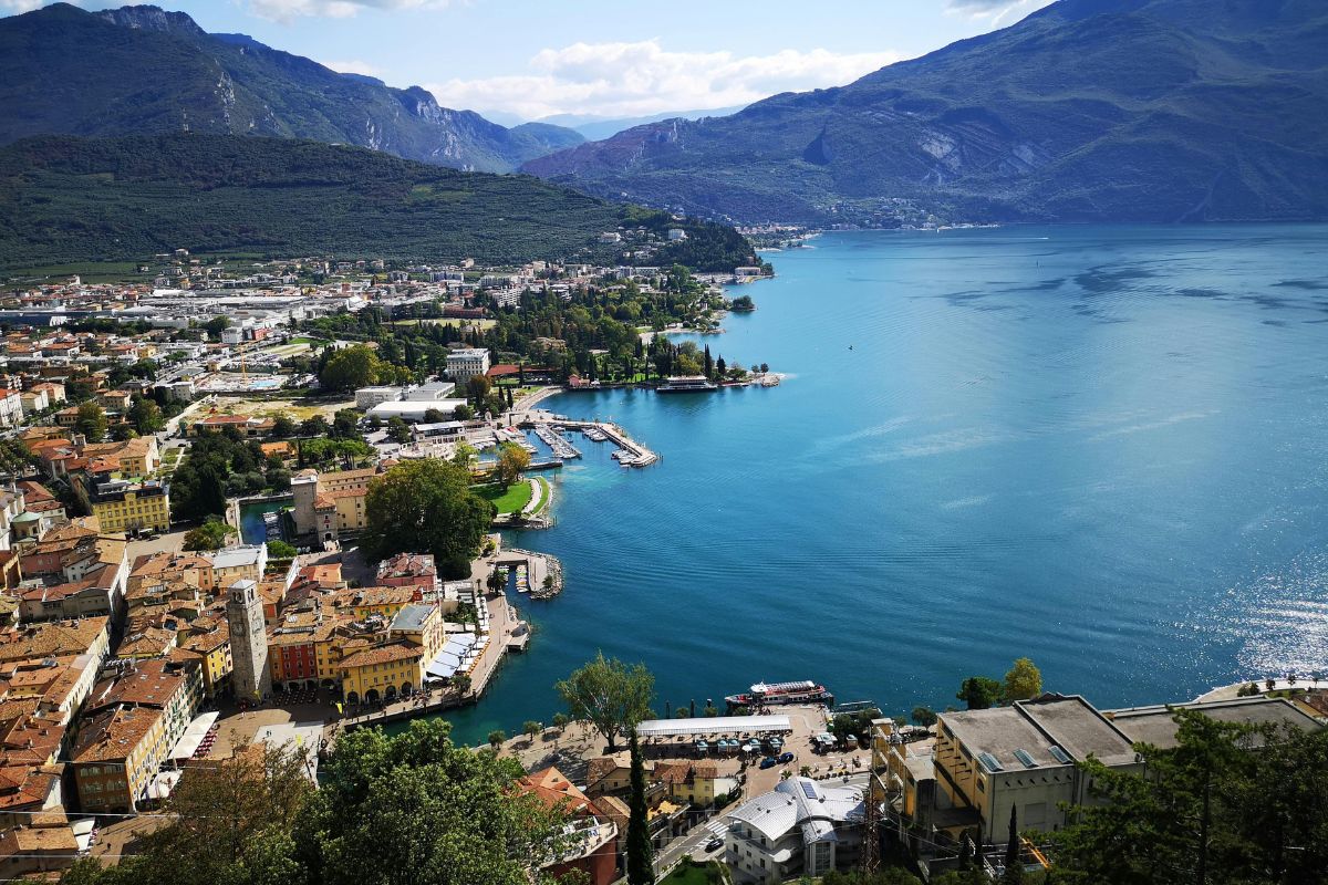 Aerial view of Riva del Garda, a town nestled on the northern shore of Lake Garda in Italy, featuring colorful buildings, a marina with boats, and surrounded by mountains. The clear blue waters of the lake contrast with the lush greenery of the landscape.