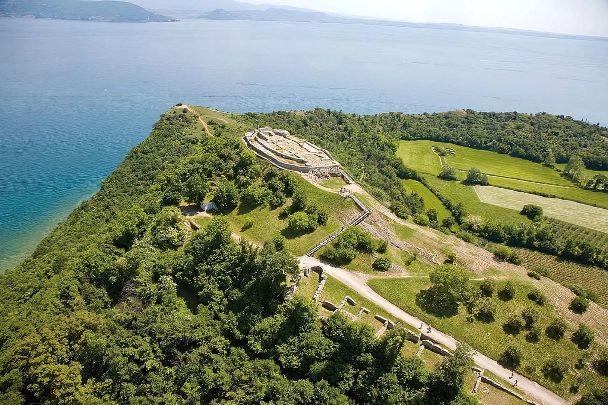 Aerial view of Rocca di Manerba, a natural reserve on a hill overlooking Lake Garda in Italy, featuring ancient ruins and lush greenery. The site includes walking paths, steps leading up the hill, and surrounding agricultural fields.
