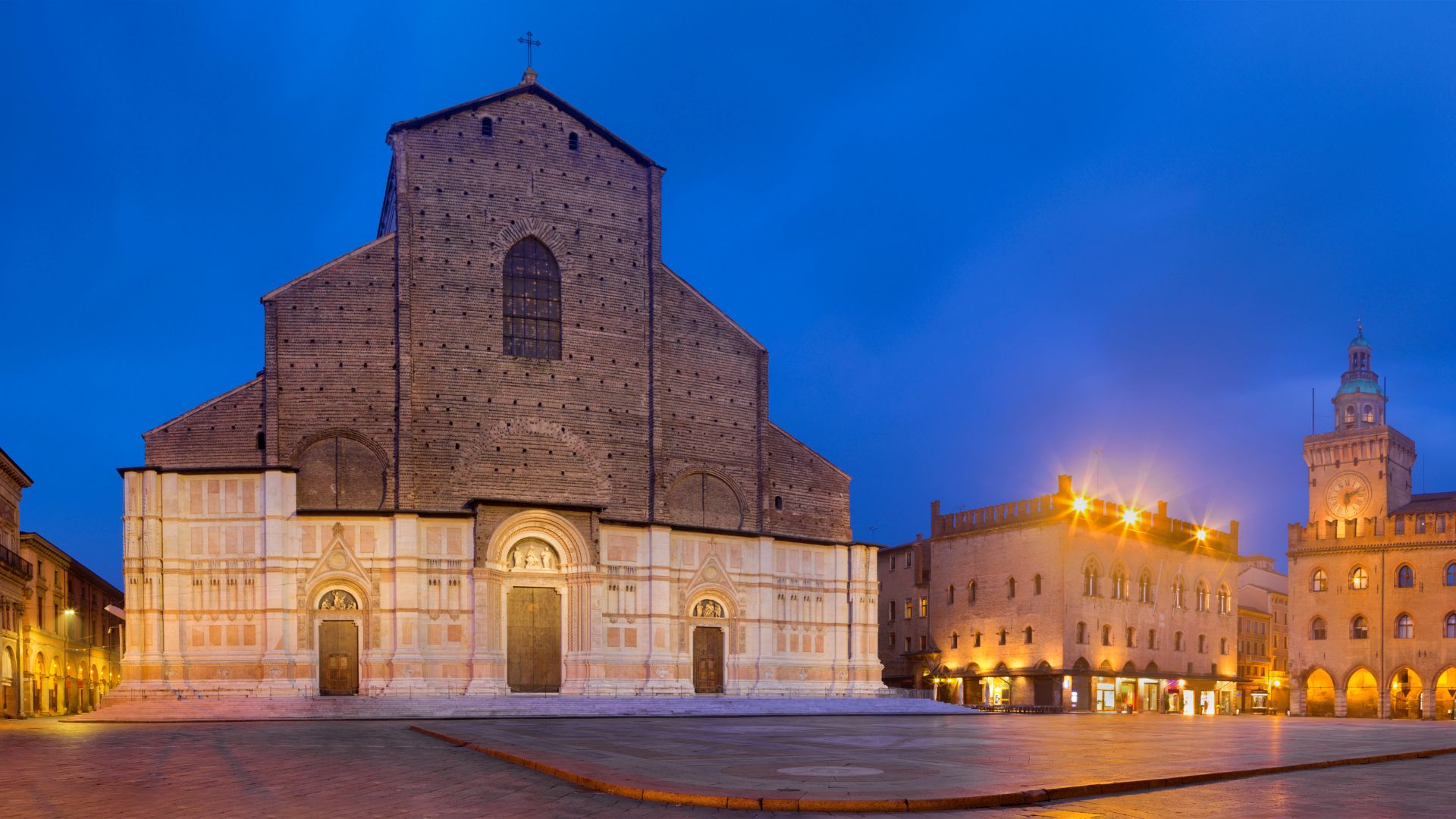 The image shows the Basilica di San Petronio in Bologna, Italy, at dusk. The basilica's facade is prominently featured, with its unfinished upper portion contrasting with the marble lower section. The Piazza Maggiore is visible in front of the basilica, and other historic buildings surround the square, including the Palazzo dei Notai
