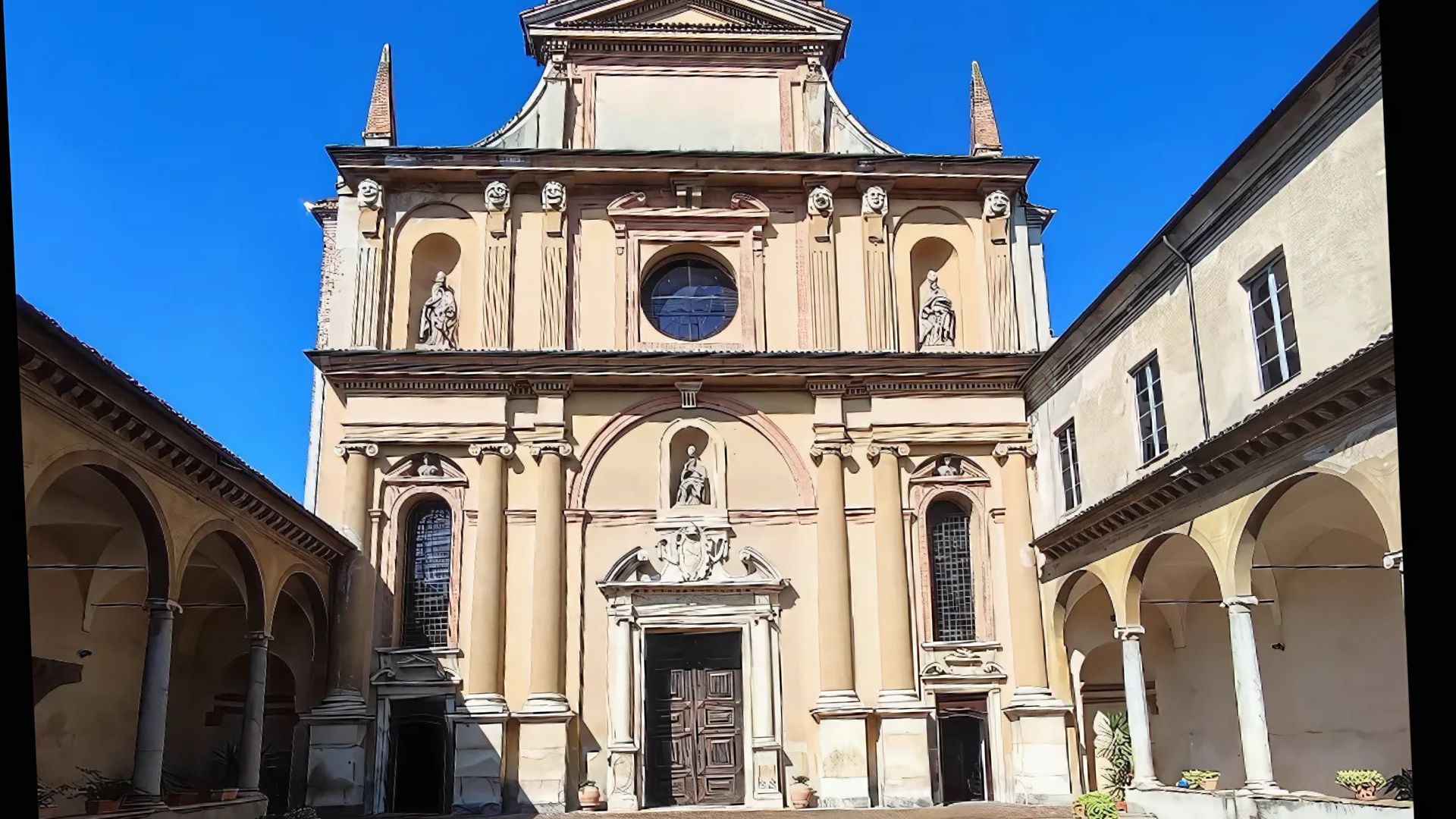 Facade of the Church of San Sisto in Piacenza, featuring a symmetrical design with a central entrance, arched windows, and statues within niches, under a clear blue sky. The church is flanked by colonnaded courtyards, showcasing its architectural details and historical presence.
