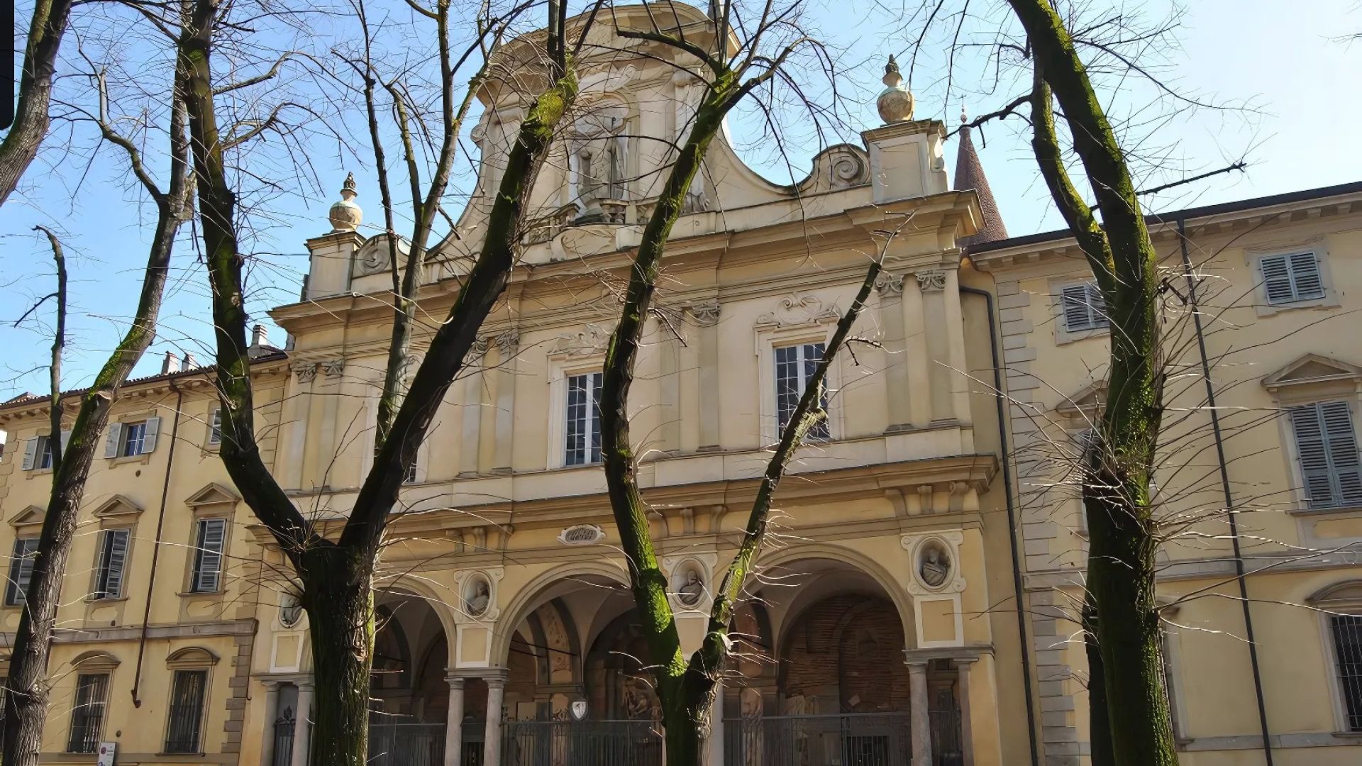 Facade of the Basilica di San Savino in Piacenza, Italy, featuring a pale yellow exterior with arched entrances and classical architectural details, partially obscured by trees in the foreground. The building exhibits a multi-tiered design with decorative elements, including statues and ornate window frames. The sky is visible in the background. The image captures the architectural style and historical ambiance of the basilica, a notable landmark in Piacenza.