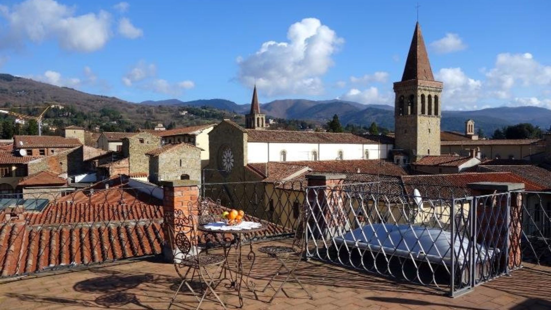 The image shows a rooftop view in Sansepolcro, Italy, specifically from the Casa Vacanze Antica Torre Medioevale d'Uguccione. The view includes the rooftops of the town, with the bell tower of the Sansepolcro Cathedral and the Apennine Mountains visible in the background.