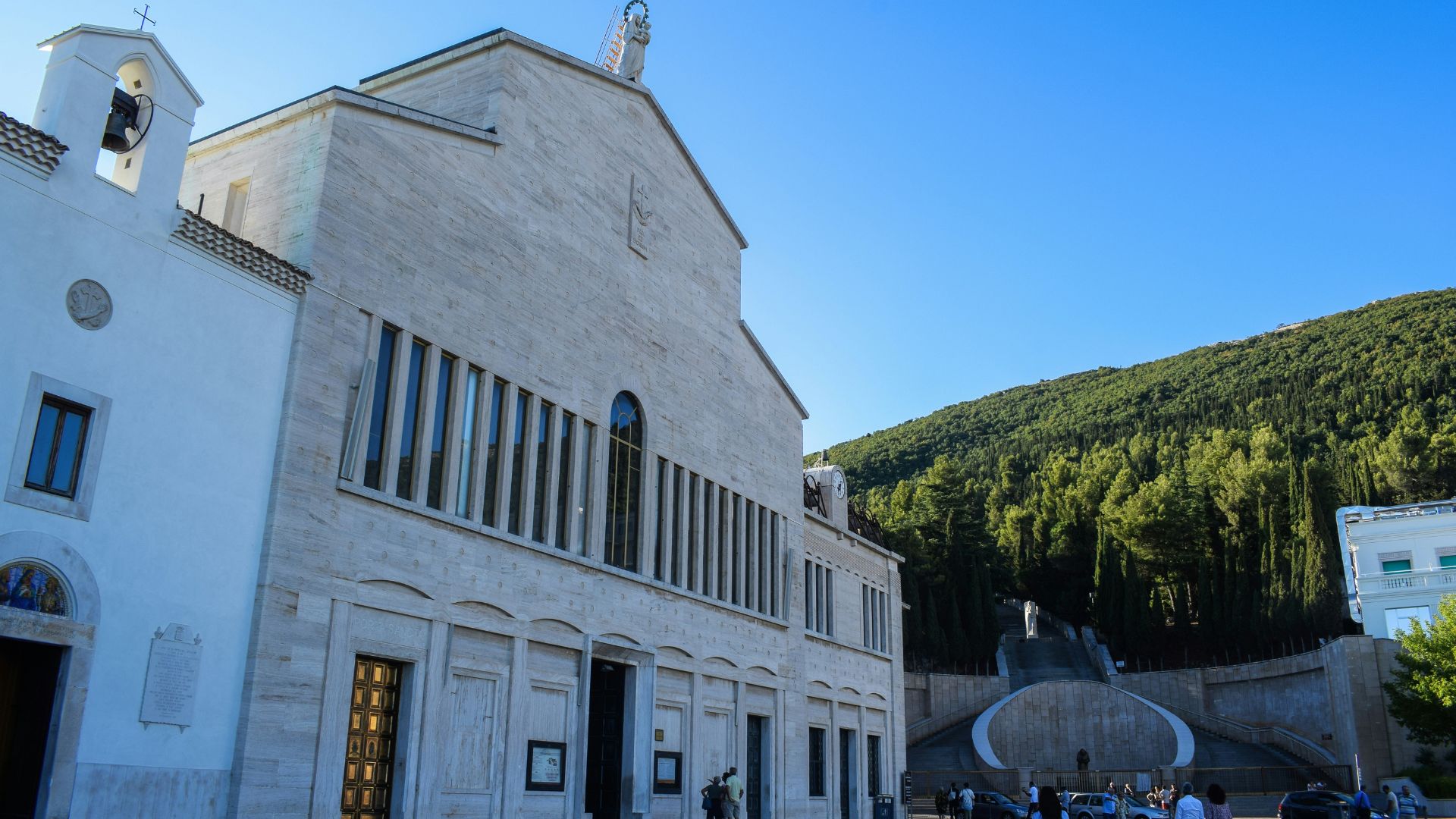 Exterior view of the Sanctuary of Saint Pio of Pietrelcina, a modern church with a light-colored stone facade, featuring a series of tall, narrow windows and a statue atop its apex, set against a clear blue sky and a backdrop of a forested hill, with people and cars visible in the foreground plaza.