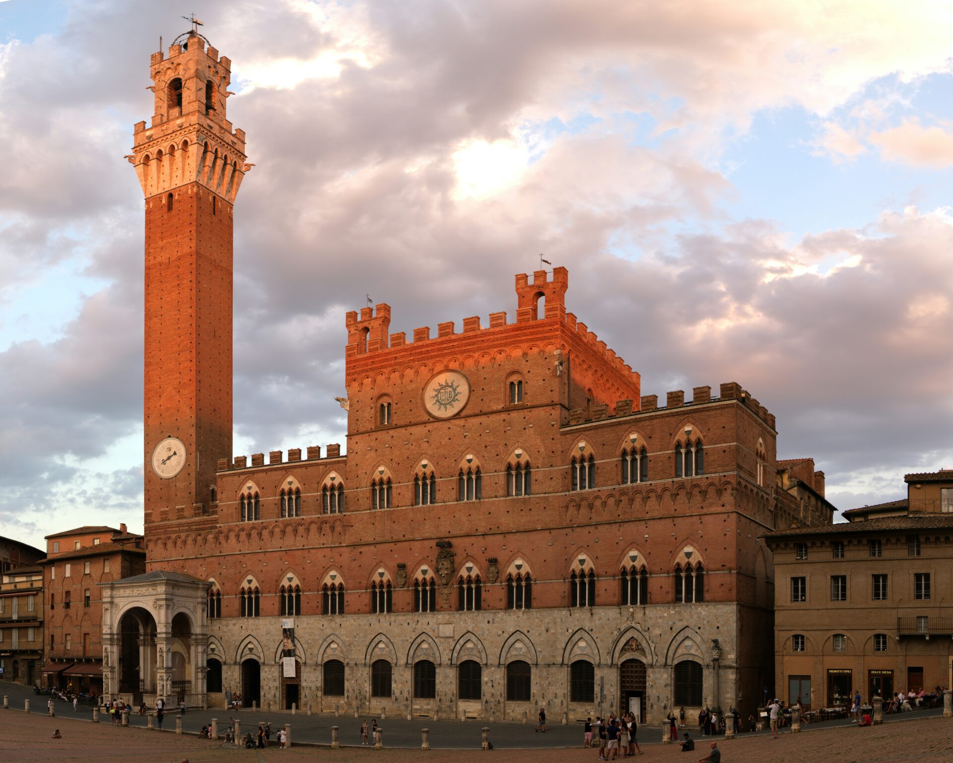 The Torre del Mangia, a tall medieval bell tower made of red brick and stone, rising above the Palazzo Pubblico in Siena, Italy, against a clear blue sky.