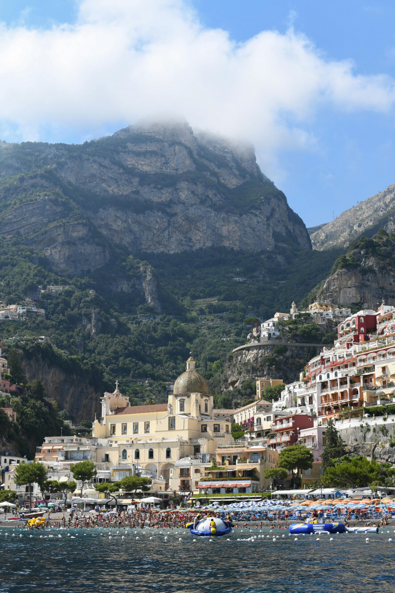 View of Spiaggia Grande beach in Positano, Italy, with colorful umbrellas, beachgoers, and the Church of Santa Maria Assunta against a mountain backdrop.