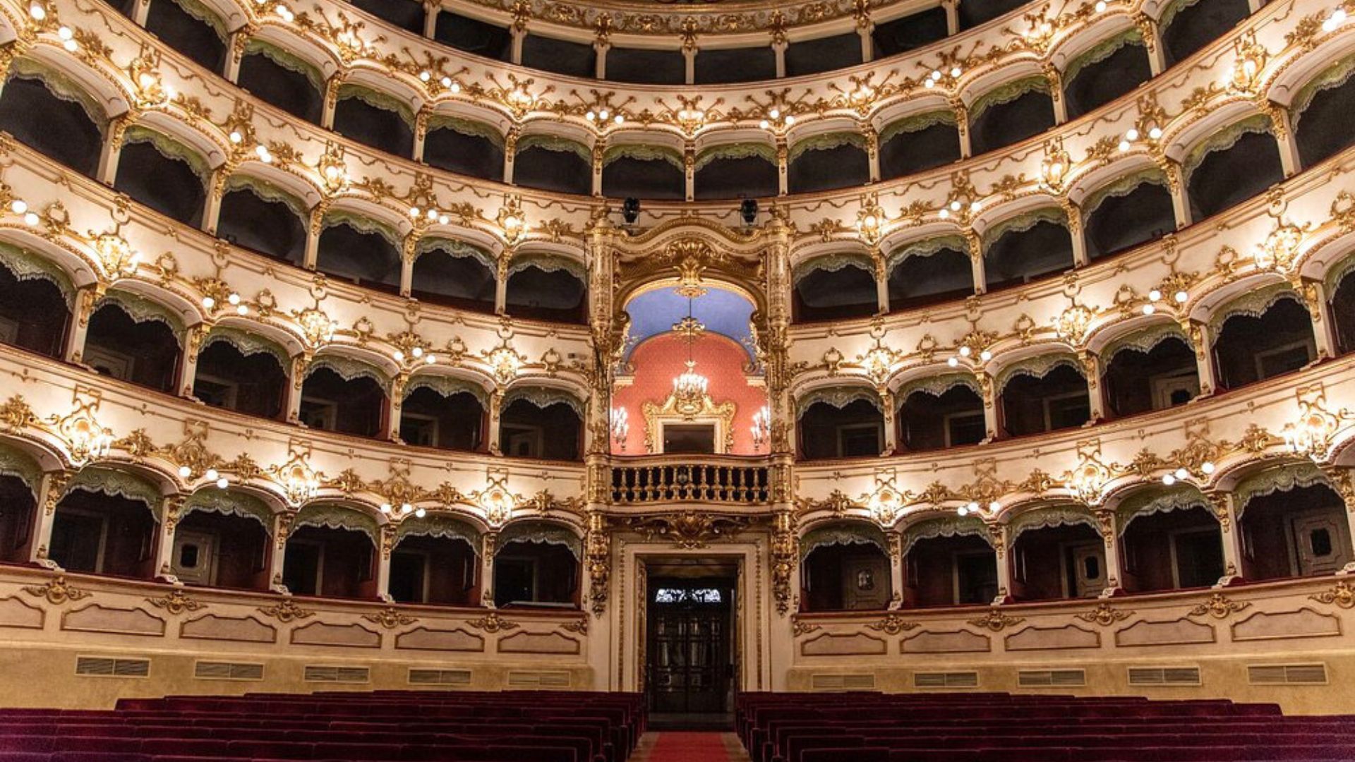 Interior view of Teatro Municipale in Piacenza, showcasing the ornate balconies and seating arrangement. The theater's design features a multi-tiered layout with detailed gold and white decorations, illuminated by soft lighting, creating a classical and elegant atmosphere. Red velvet seats are arranged in rows on the ground level, facing the stage.
