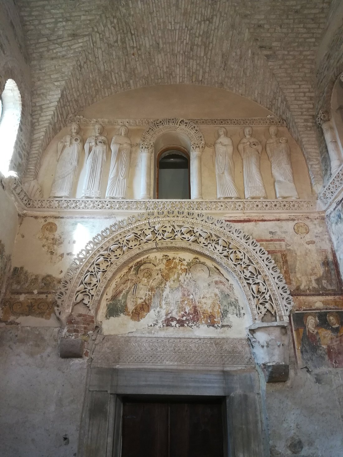  Interior view of the Tempietto Longobardo in Cividale del Friuli, Italy, showing detailed stone carvings, arched frescoes, and a row of elegant statues above a decorative doorway.