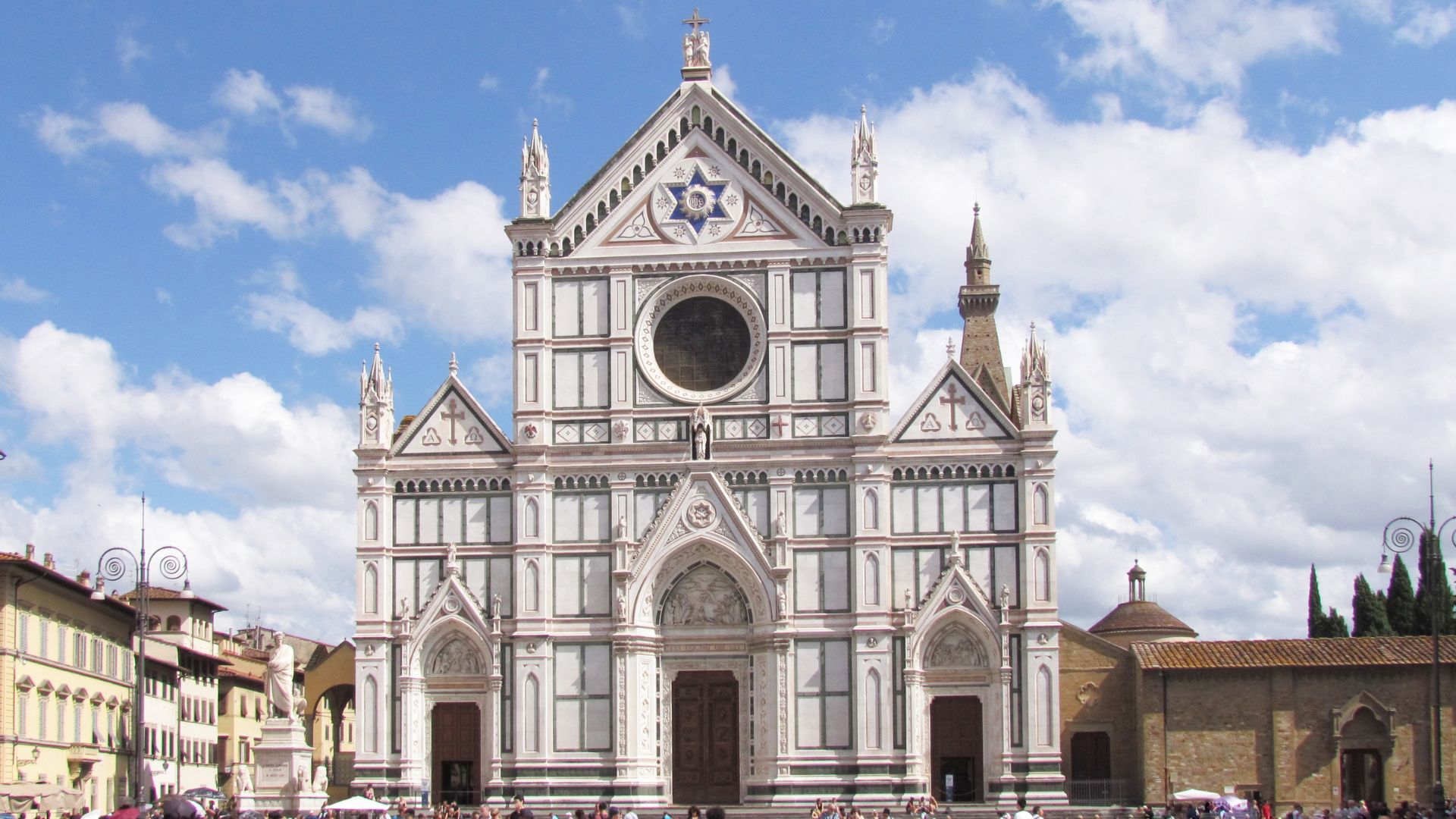 The Basilica di Santa Croce, a large Franciscan church in Florence, Italy, is depicted with its distinctive facade of white marble panels, pointed arches, and decorative elements. The building's structure features a central rose window, a large arched entrance, and several smaller entrances, all contributing to its elaborate Gothic Revival style. 