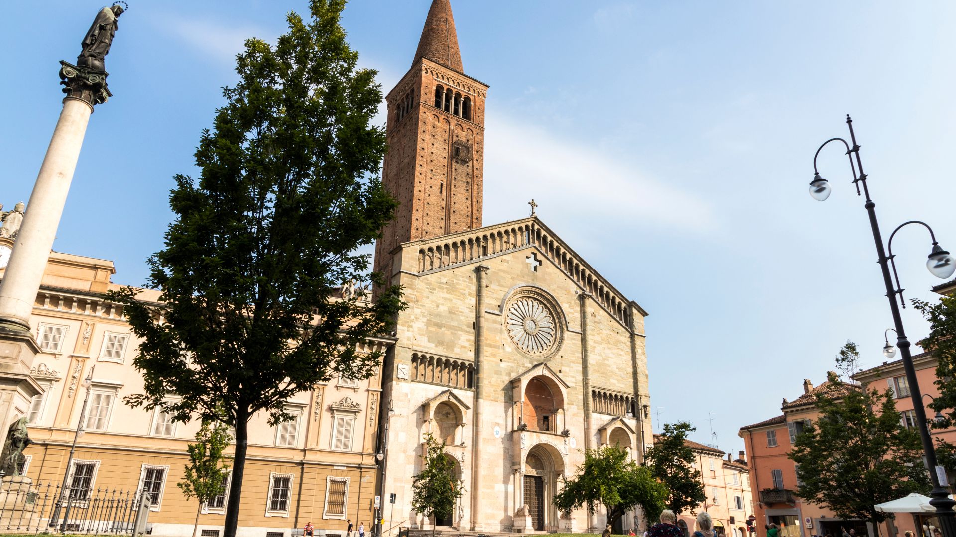 Piacenza Cathedral, featuring a Romanesque facade with a rose window and a tall brick bell tower, stands prominently in a city square with surrounding buildings and trees under a clear sky.
