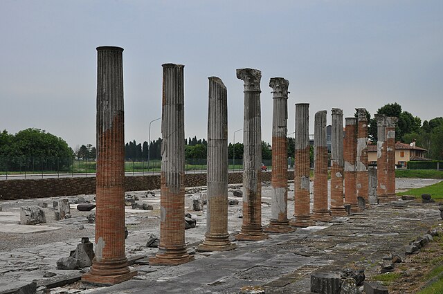 Ancient stone and brick columns of the Roman Forum of Aquileia, Italy, standing in a row on a historic archaeological site under a cloudy sky.