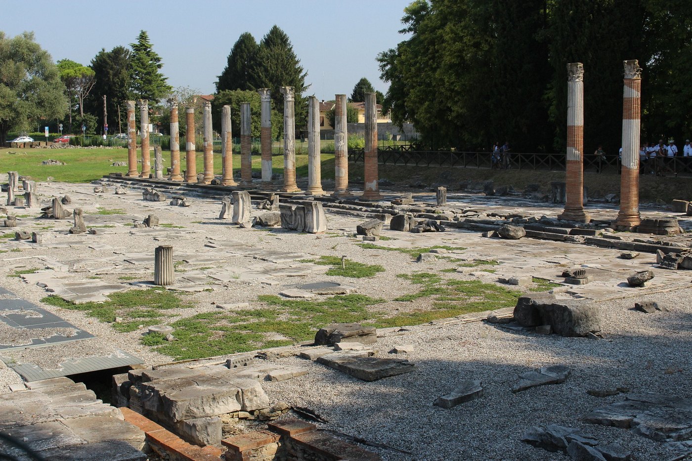 Ancient stone columns and ruins at the Roman Forum in Aquileia, Italy, with scattered remnants of Roman architecture set in an open grassy area surrounded by trees.