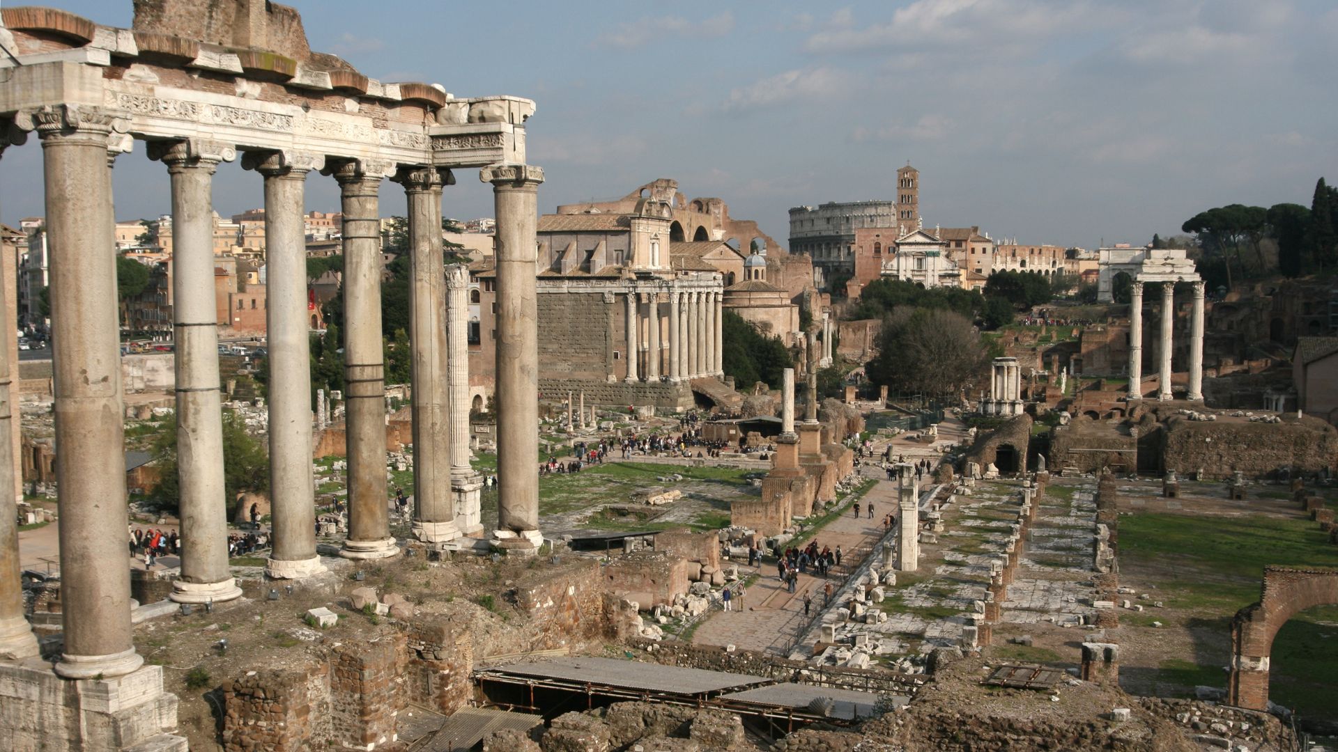 The image shows a partial view of the Roman Forum, including columns and ruins, with people walking along pathways. The Via Sacra, or Sacred Way, was the main street of ancient Rome, leading from the Capitoline Hill through the Forum to the Colosseum.