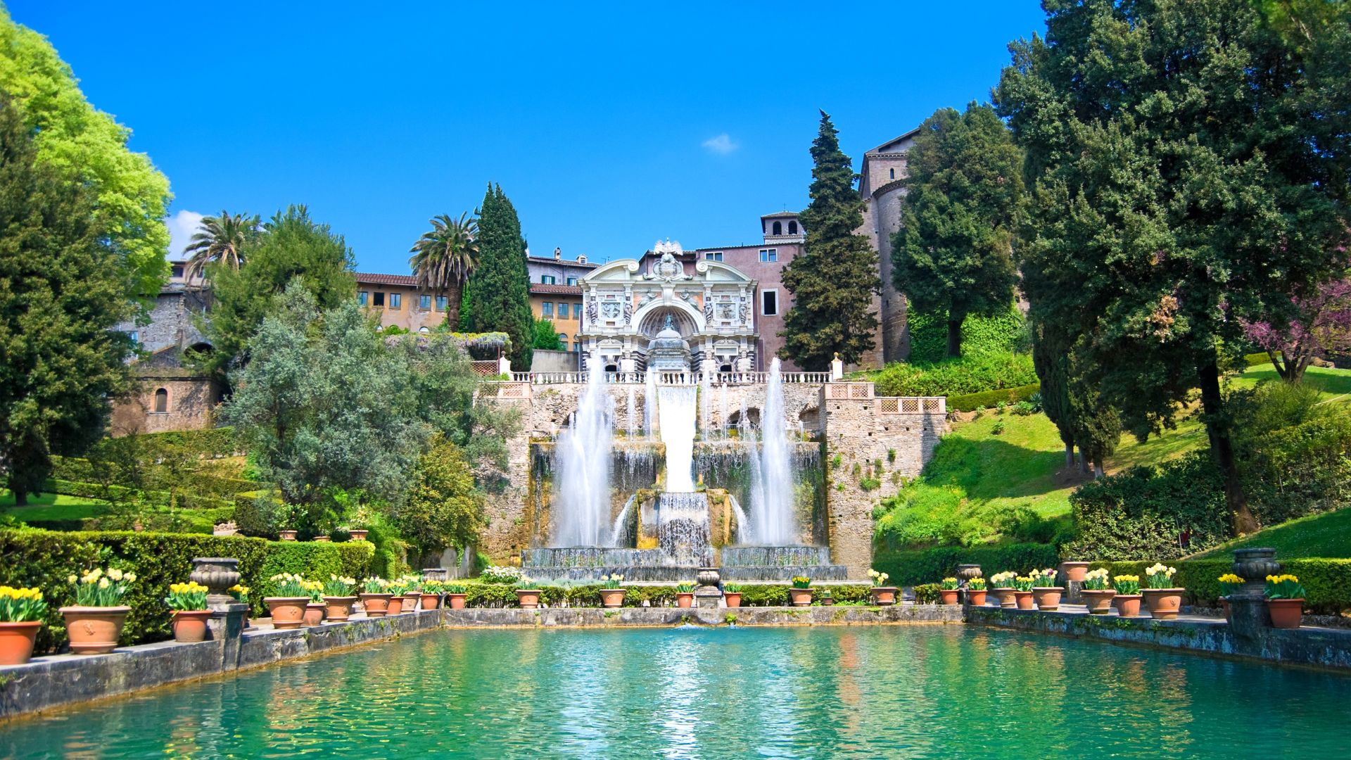 The image of Tivoli's Villa d'Este shows a large fountain complex with multiple cascades flowing into a pool lined with potted plants. The villa itself is visible in the background, set against a backdrop of trees and a clear blue sky.