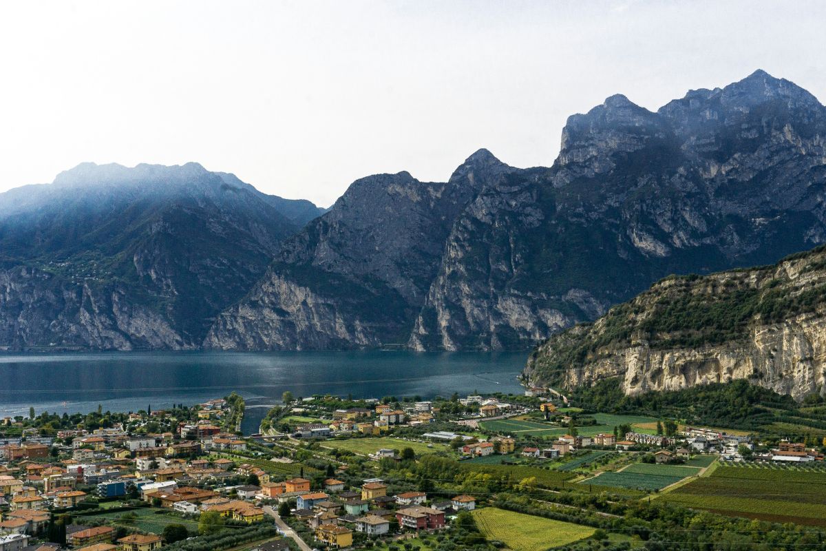 Torbole, a town on the northern shore of Lake Garda, Italy, is nestled at the foot of the Alps, with its buildings clustered near the water and surrounded by greenery. The town is set against a backdrop of rugged mountains that rise dramatically from the lake's edge