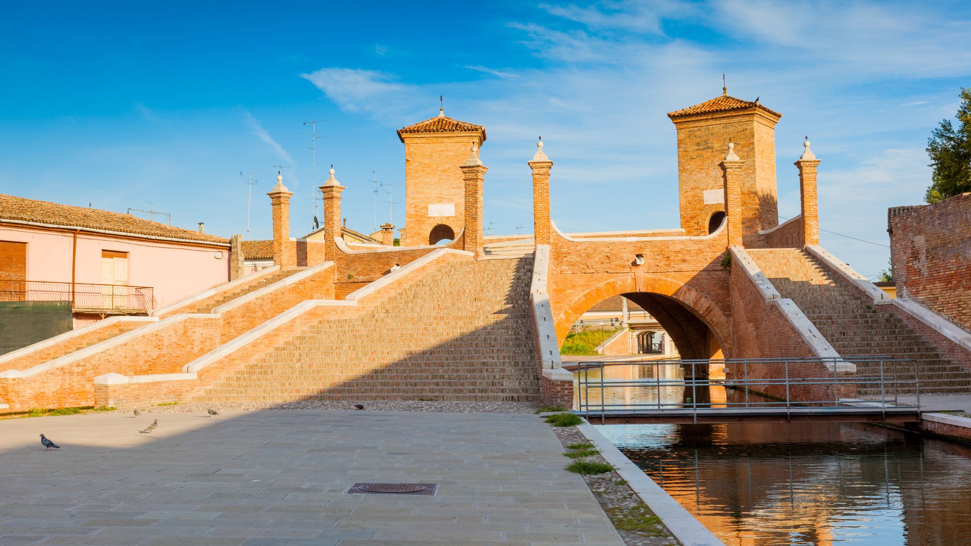 Ponte dei Trepponti, a historic brick bridge with two towers and staircases, spans a canal in Comacchio, Italy, under a clear blue sky. It is a famous landmark and symbol of the town.