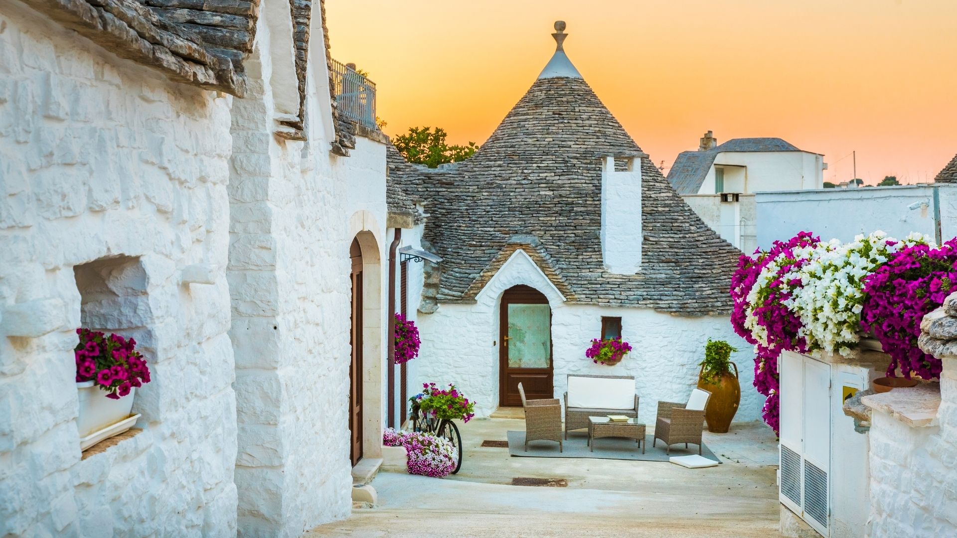 The image shows a row of Trulli houses in Alberobello, Italy. These traditional Apulian dry stone dwellings have distinctive conical roofs, some adorned with symbolic signs. The houses are whitewashed with dark wooden doors, and small plants and flower boxes decorate the entrances. The street in front is paved, and the sky is clear and blue. The scene captures the unique architectural style and charm of the Itria Valley region.