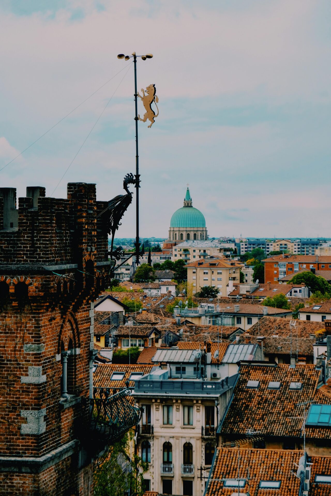 Scenic view of Udine, Friuli-Venezia Giulia, Italy, featuring terracotta rooftops, historic buildings, and the green dome of a prominent church in the distance under a cloudy sky.
