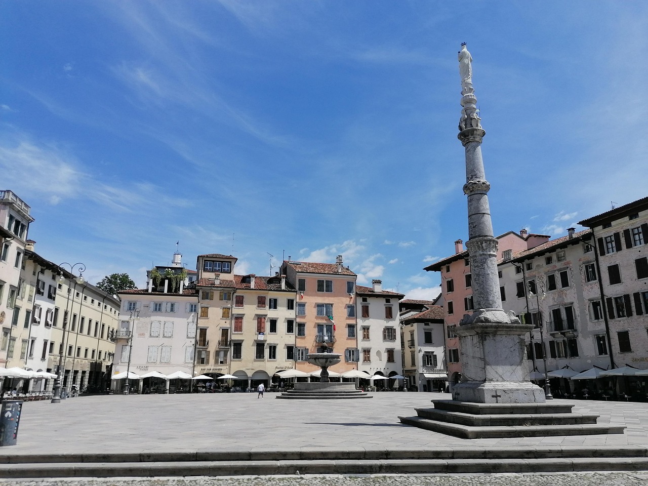 Sunny day at a historic square in Udine, Italy, with colorful old buildings, outdoor cafes, and a tall marble column topped with a statue.