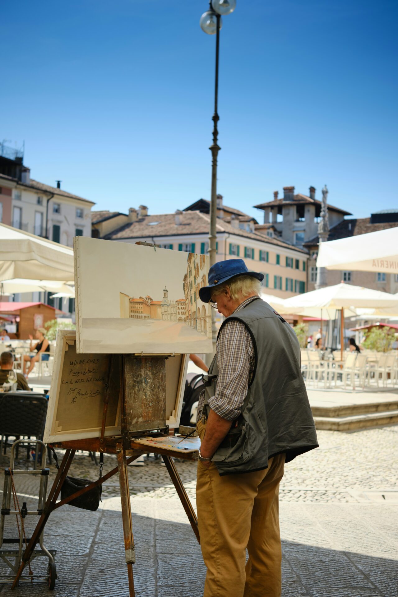 Elderly man painting a cityscape on an easel in a sunny outdoor square in Udine, Italy, surrounded by historic buildings, café tables, and umbrellas.