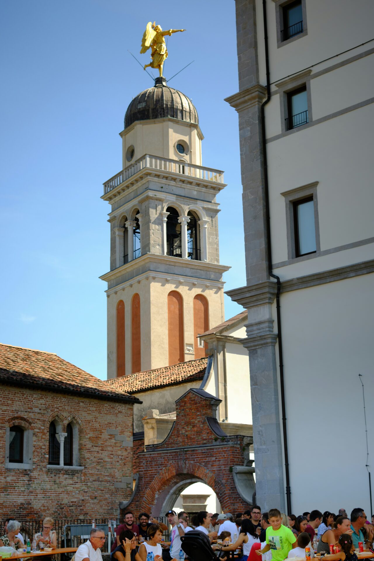 People enjoying an outdoor gathering near a historic bell tower topped with a golden angel statue in the Province of Udine, Italy, on a sunny day.