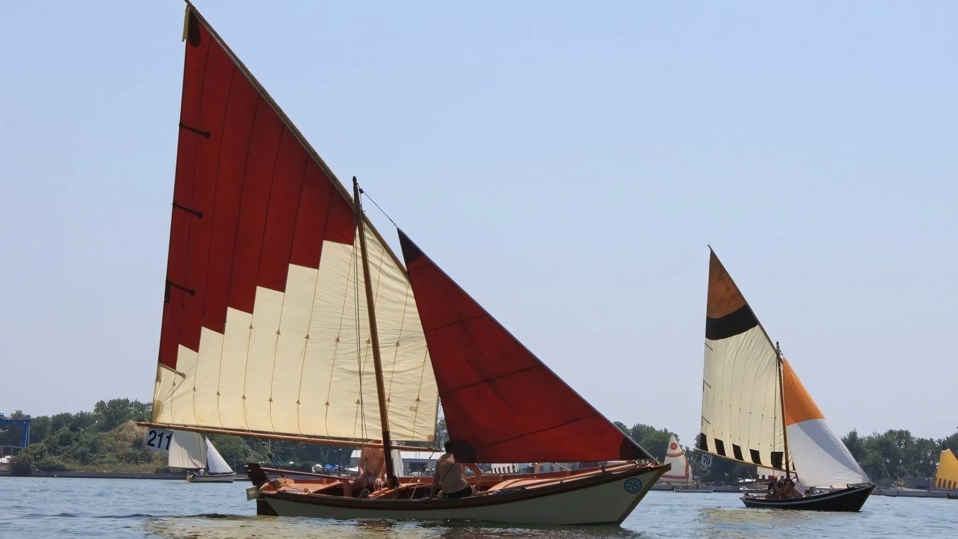 Three Vela al terzo sailboats with red and white sails are sailing on the water on a sunny day. 