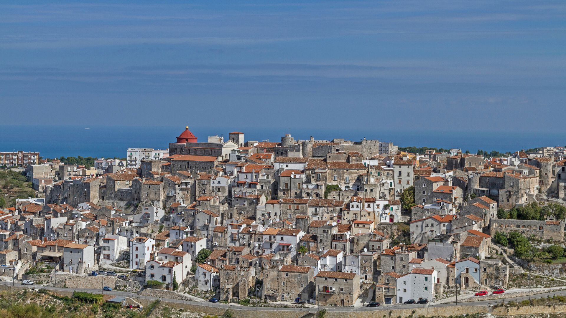 Vico del Gargano, a hilltop town in the Gargano National Park, Italy, is captured in a wide shot showcasing its dense cluster of buildings with red-tiled roofs and stone facades, set against a backdrop of the Adriatic Sea. The town's architecture, characterized by narrow streets and closely packed structures, is visible, with a church featuring a red dome standing out among the buildings.