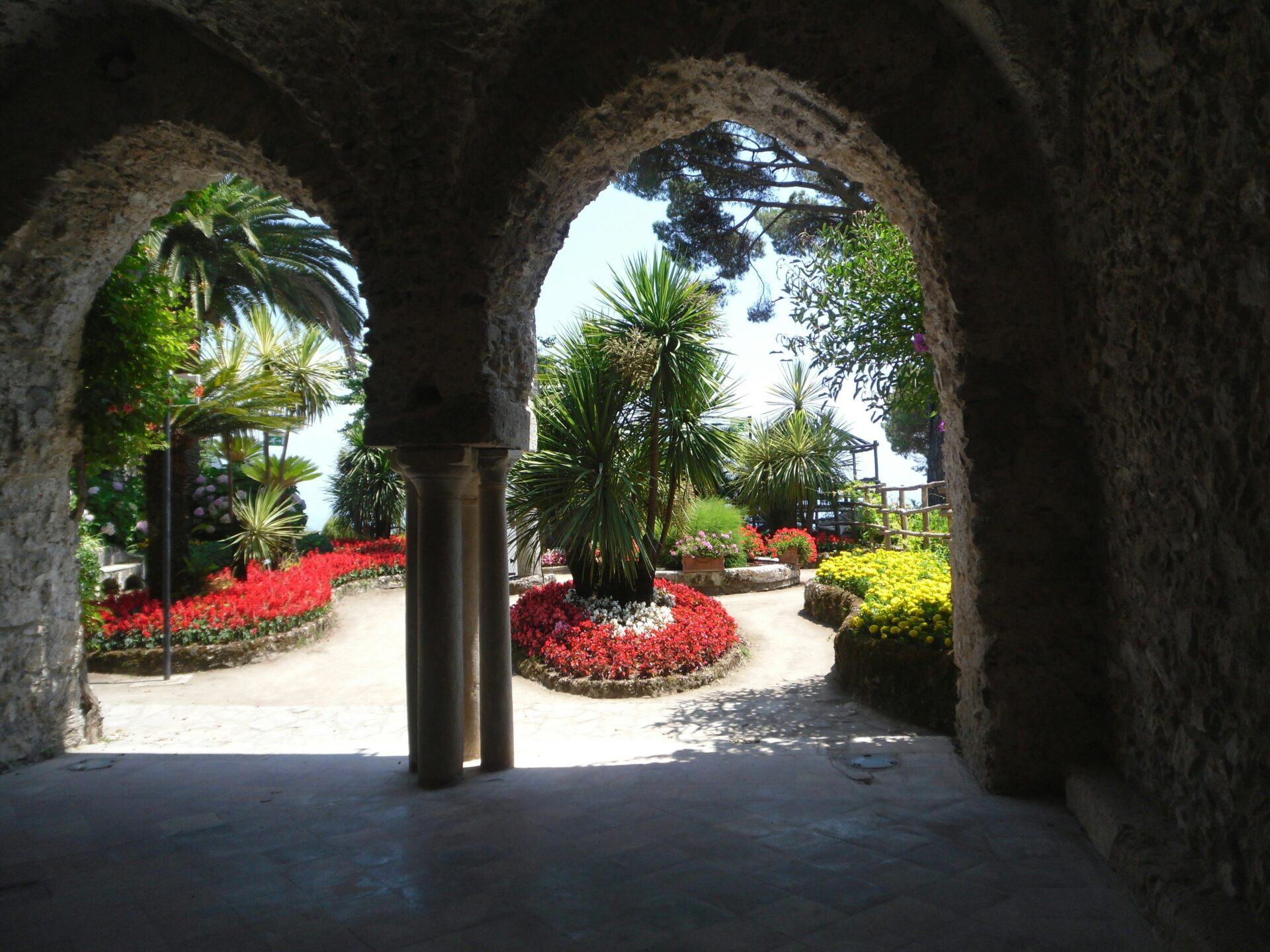 Colorful garden with palm trees and flowers seen through stone arches at Villa Rufolo in Ravello, Italy.