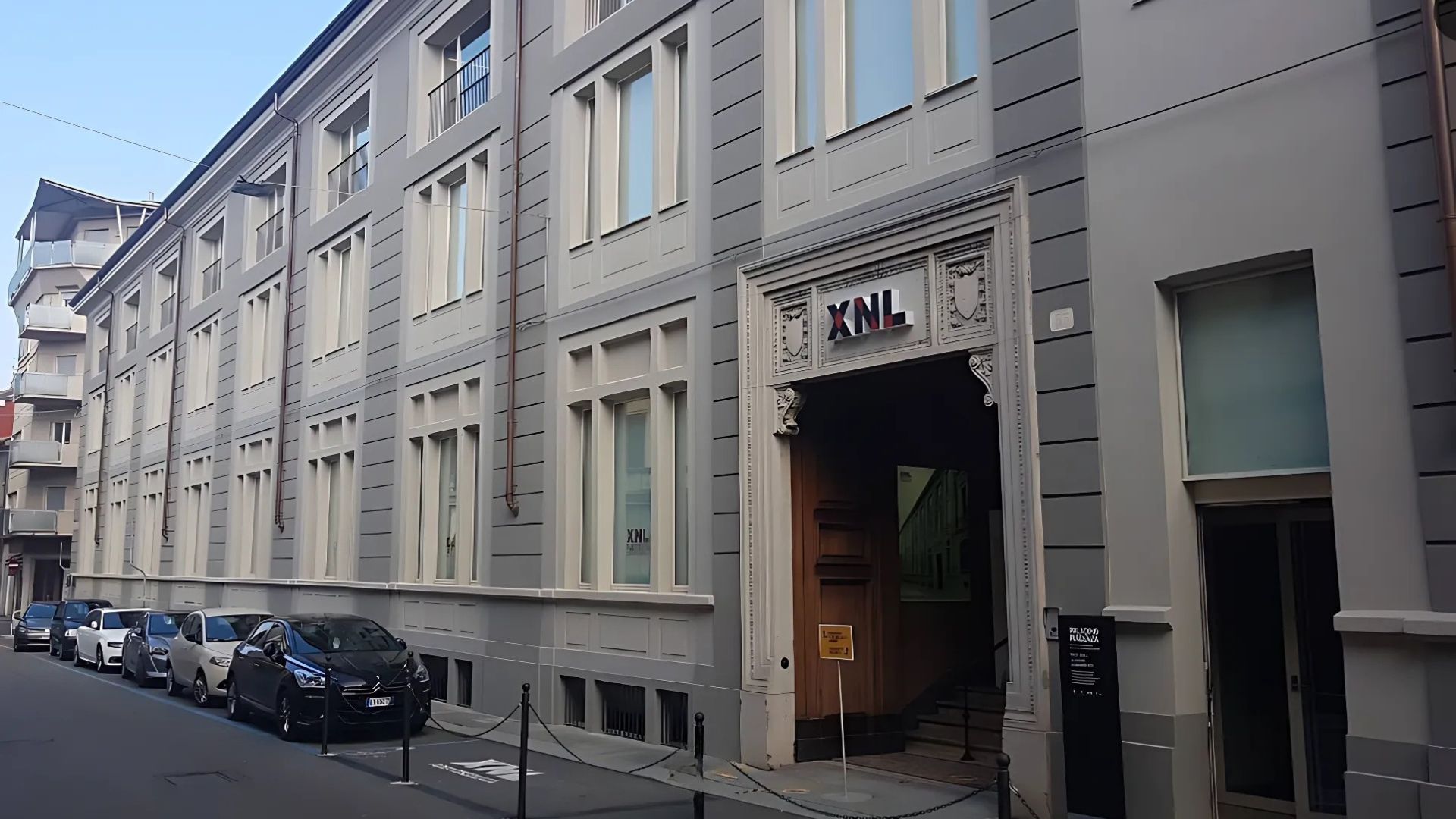 XNL Piacenza building with "XNL" sign above the entrance, captured on a street view. The building is painted in shades of gray with white window frames. Several cars are parked along the street in front of the building.