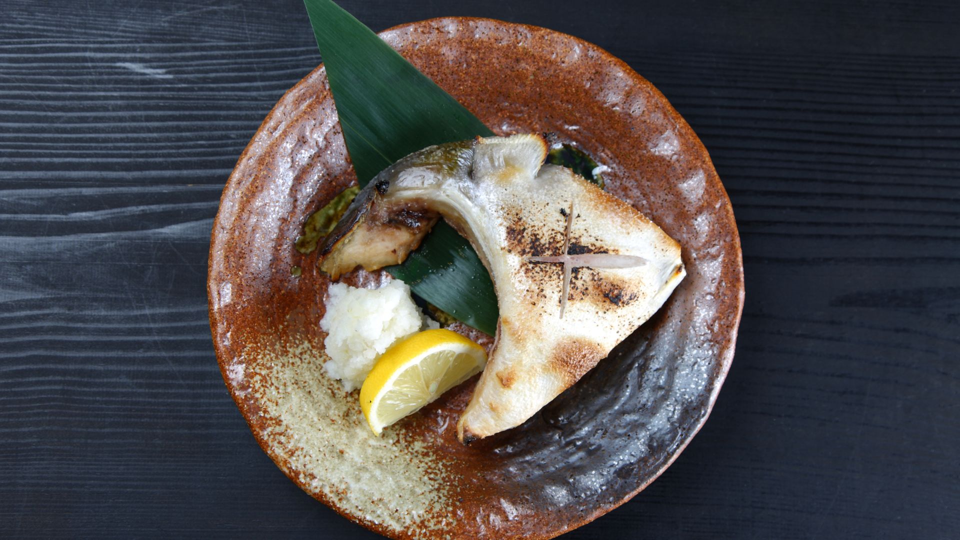 The image shows a Grilled Yellowtail Collar, which is a cut of yellowtail that is seared to preserve its buttery texture. The sear marks on the fish indicate that it was cooked quickly at high heat, which helps to keep the inside moist and tender.