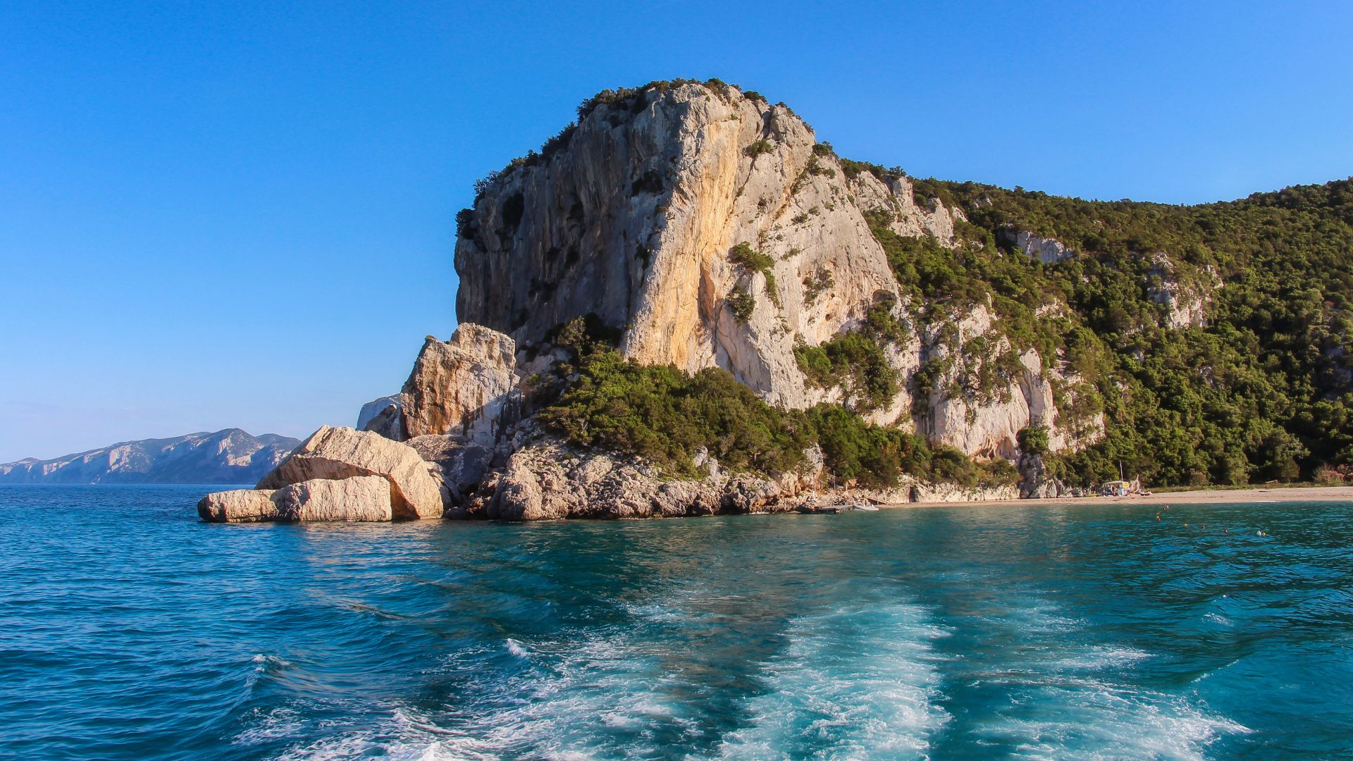 A scenic view of Cala Luna in Sardinia, featuring a sandy beach bordered by towering limestone cliffs, lush green vegetation, and the clear blue waters of the Mediterranean Sea.