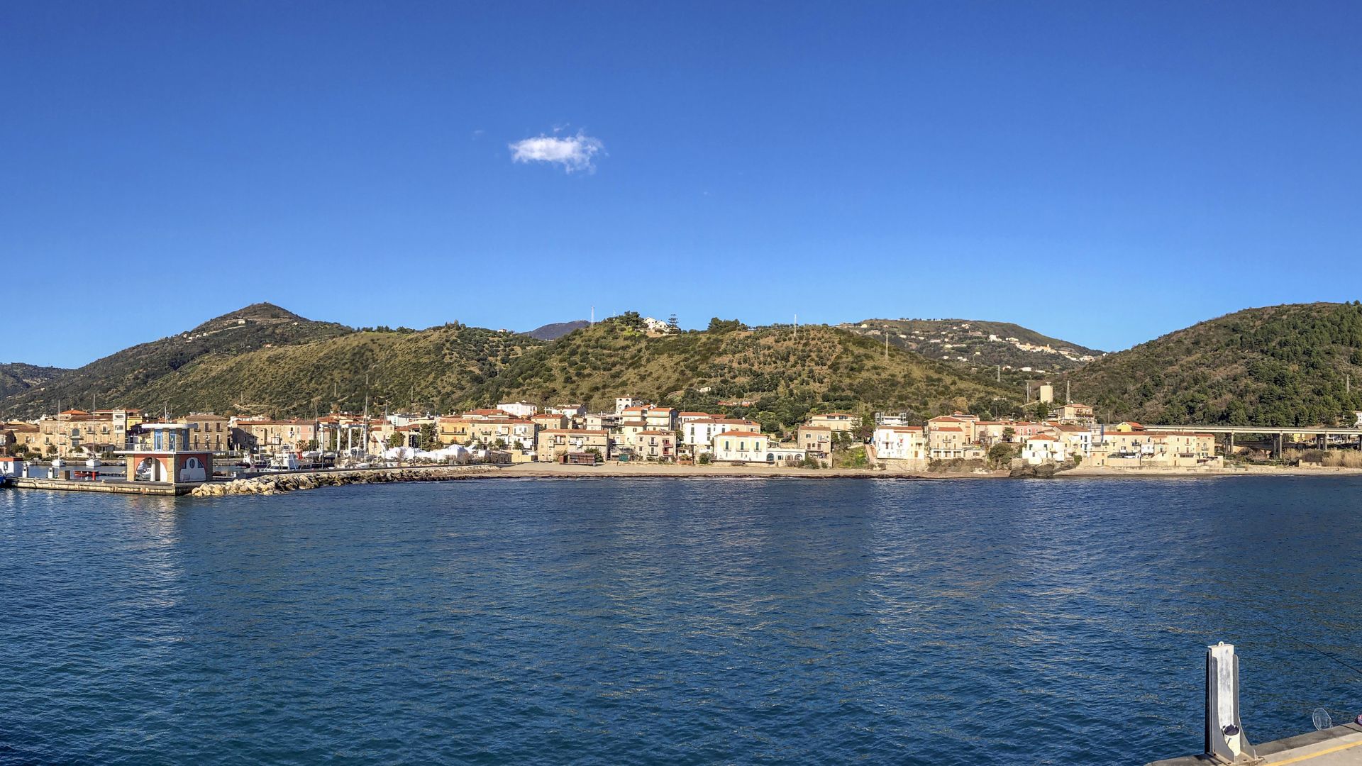 A panoramic view of Acciaroli, Italy, featuring the town's harbor, waterfront buildings, and the surrounding green hills under a clear blue sky.