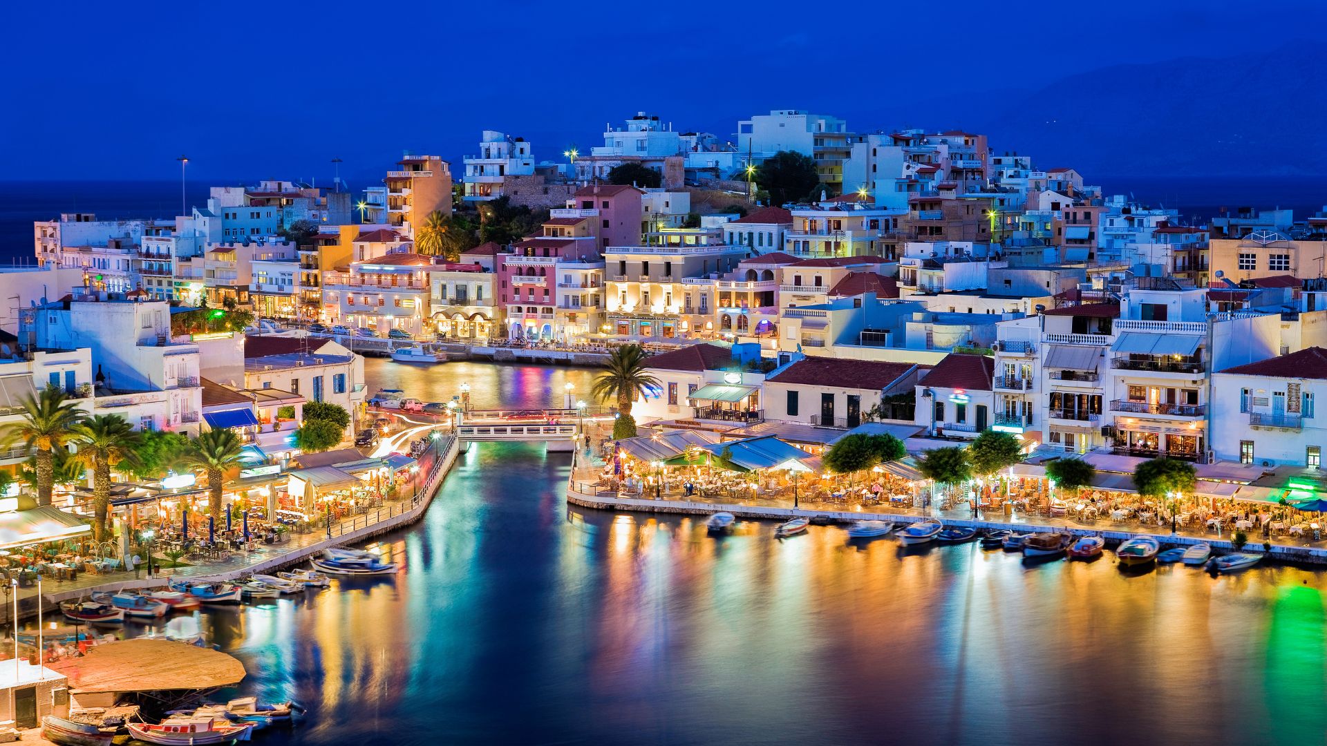 A scenic view of Lake Voulismeni surrounded by buildings and vegetation in Agios Nikolaos, Crete at night.