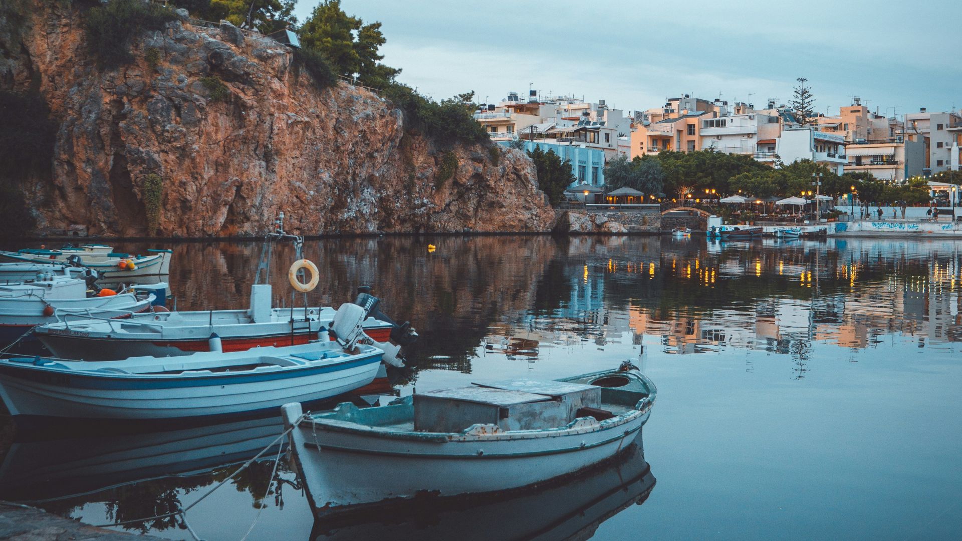 Boats moored on the calm waters of Lake Voulismeni in Agios Nikolaos, Crete, with a rocky cliff and town buildings in the background.