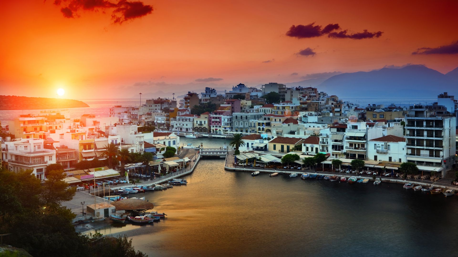 A scenic view of Lake Voulismeni in Agios Nikolaos, Crete, at sunset, surrounded by buildings and boats.