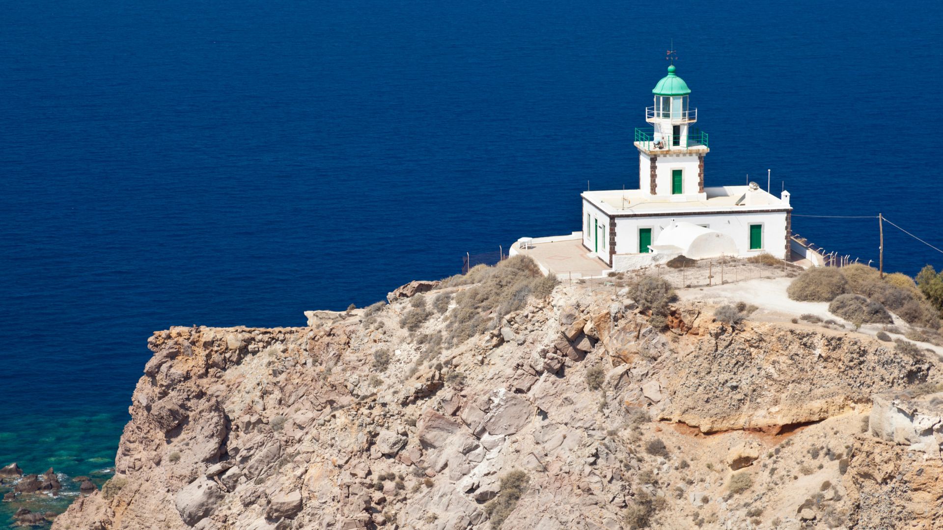 A white lighthouse sits atop a rocky cliff overlooking the blue sea on a sunny day.