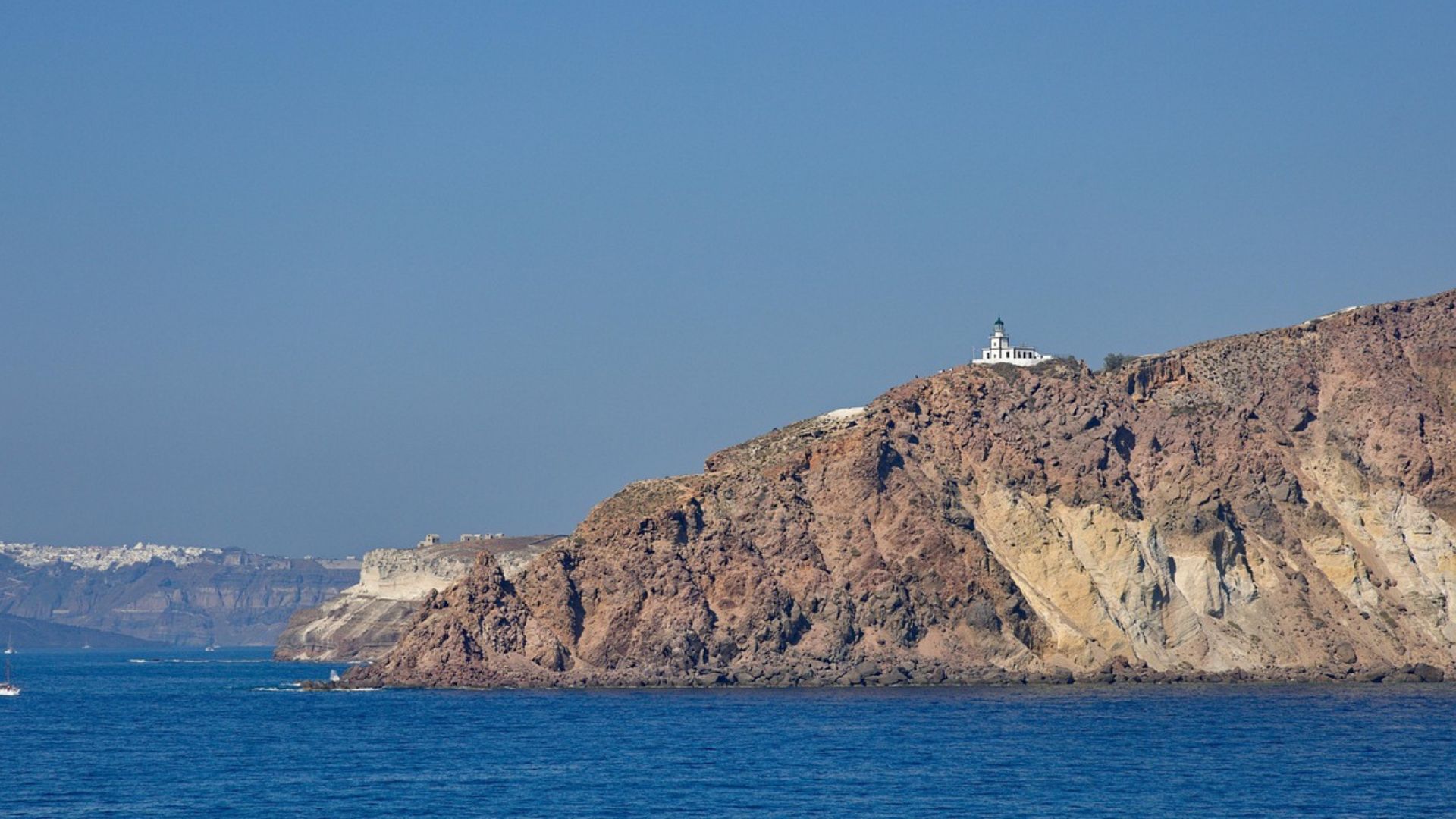 A white lighthouse sits atop a rocky cliff overlooking the blue sea on a sunny day.