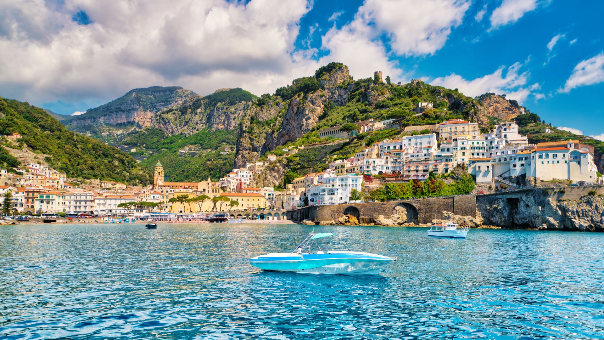 Scenic view of clear blue waters, green mountains and buildings at the back, and boats. 