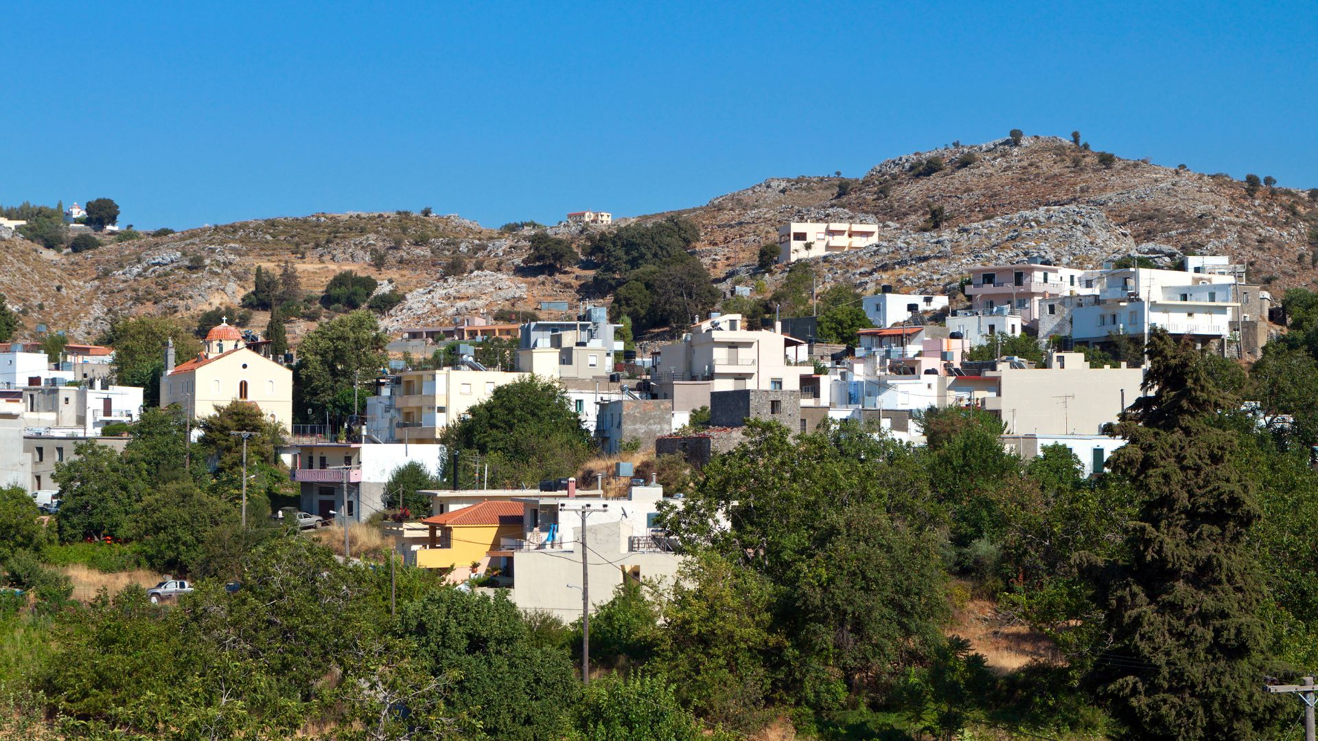 Picturesque view of Anogia village nestled on the slopes of Mount Psiloritis in Crete, featuring white houses, green trees, and a clear blue sky.