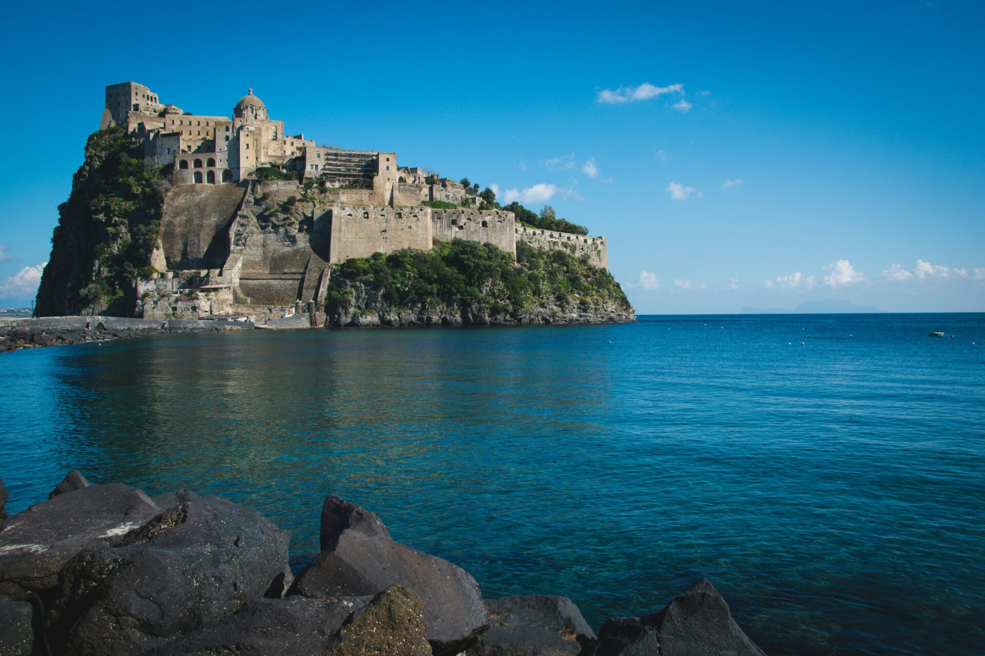 Distant daytime view of the Aragonese Castle rising from the coastline, surrounded by blue sea and set against a clear sky.