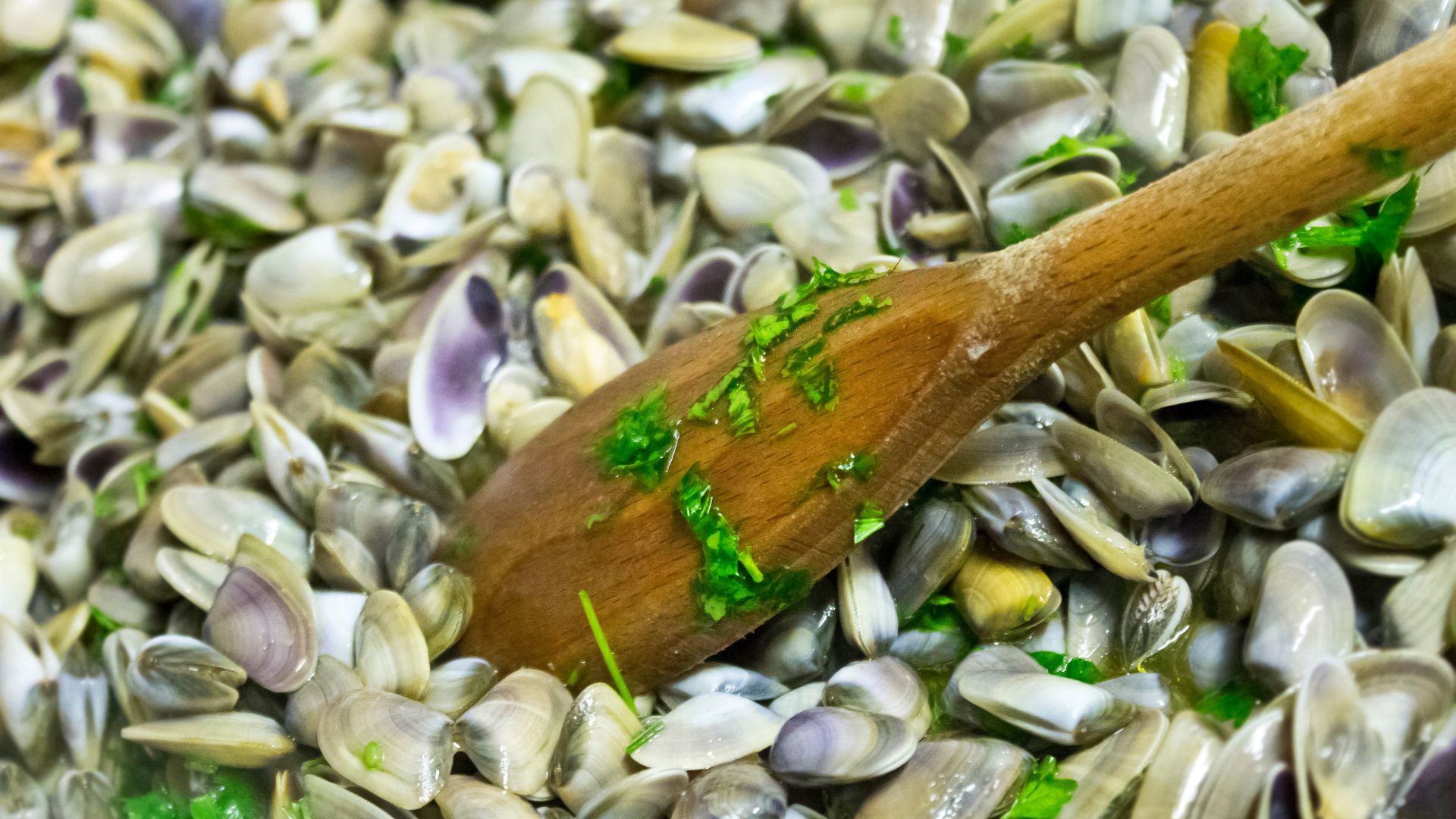 Pan filled with small clams, some of which have opened, and a wooden spoon resting on top.