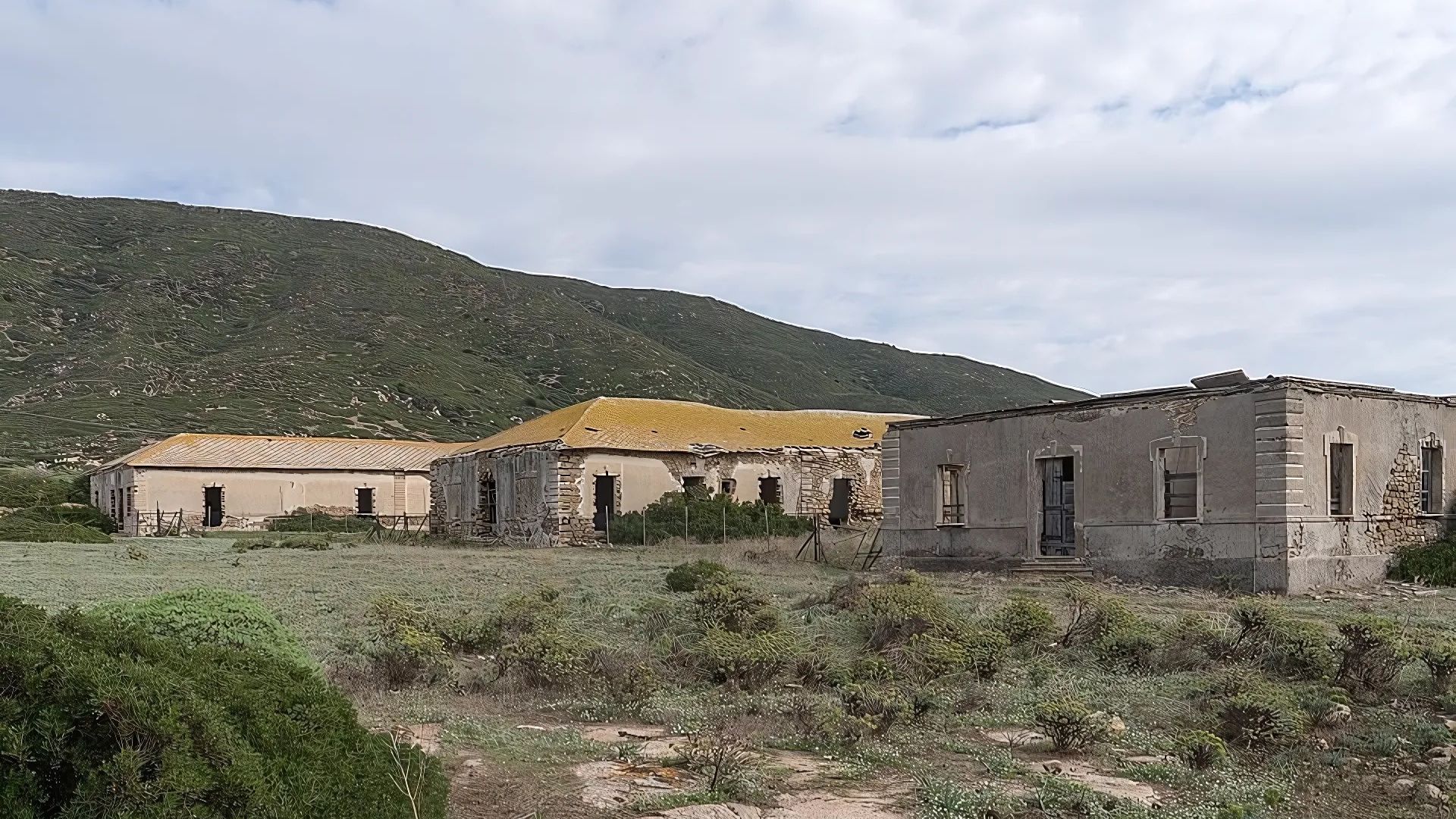 Three weathered, abandoned buildings stand in a grassy field, with a green, hilly landscape in the background under a cloudy sky. The buildings show signs of disrepair, with missing roofs and crumbling walls, suggesting they are remnants of the island's past.