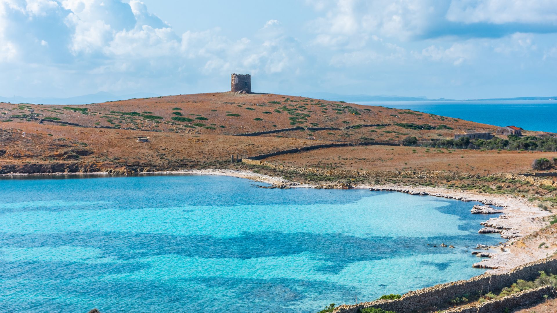 A scenic view of Asinara Island features a bay with clear turquoise water in the foreground, a coastline with a stone wall, and a hill topped with the Trabuccato Tower in the background, under a partly cloudy sky.