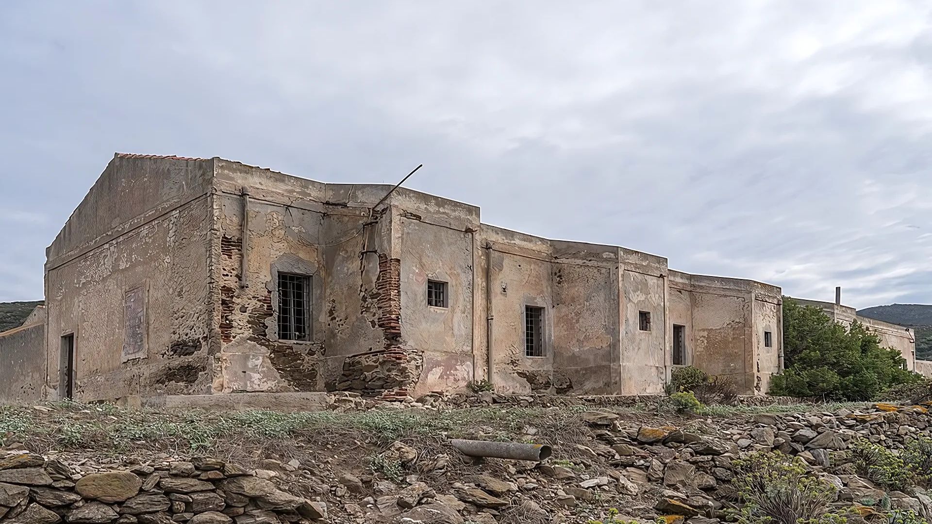 An old, weathered building with crumbling walls and barred windows stands against a cloudy sky, set amidst a rocky landscape with sparse vegetation on Asinara Island.