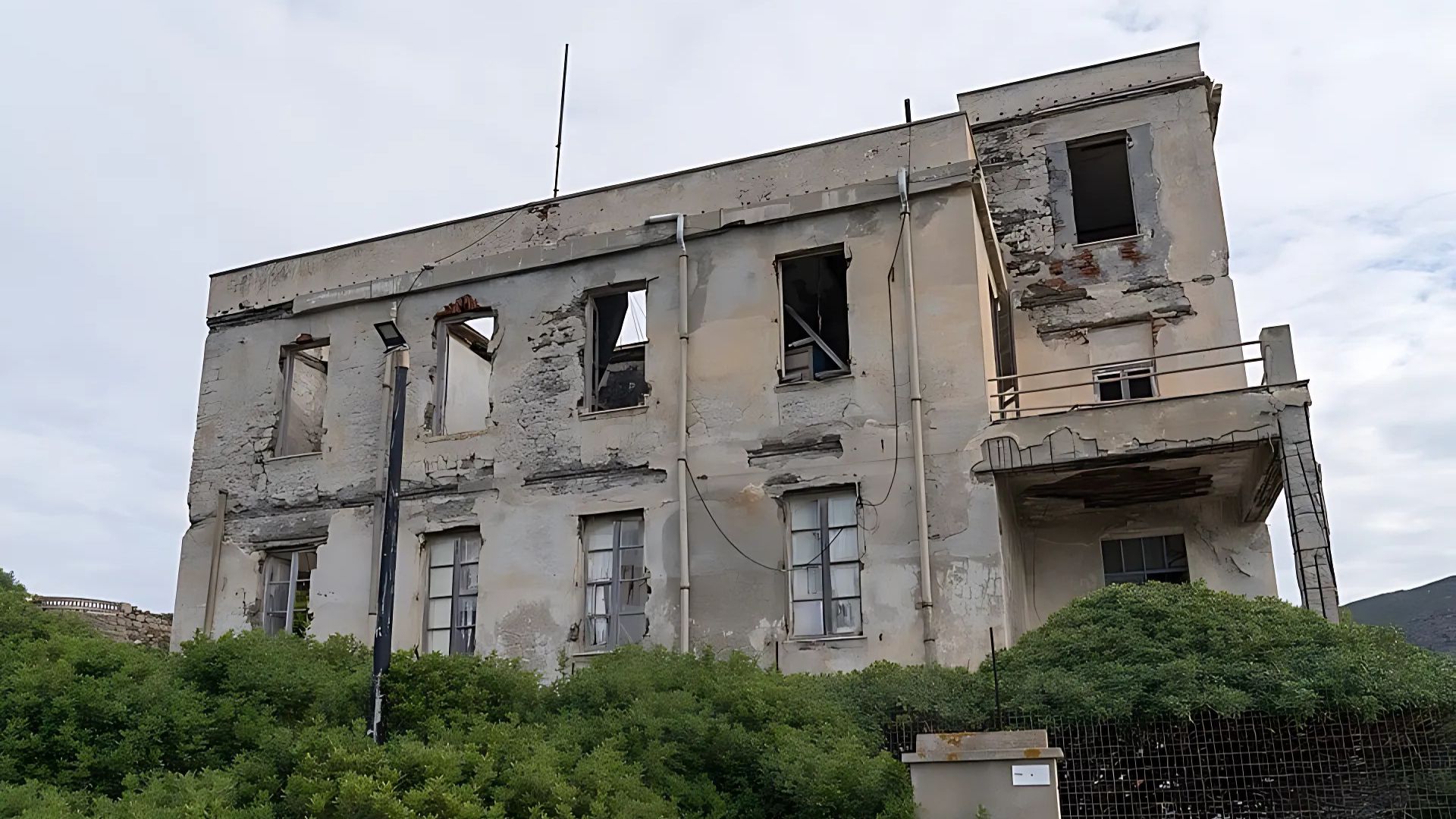 A distressed, old building with broken windows and peeling paint, possibly the former Alcatraz prison, is partially obscured by lush green bushes under a cloudy sky.
