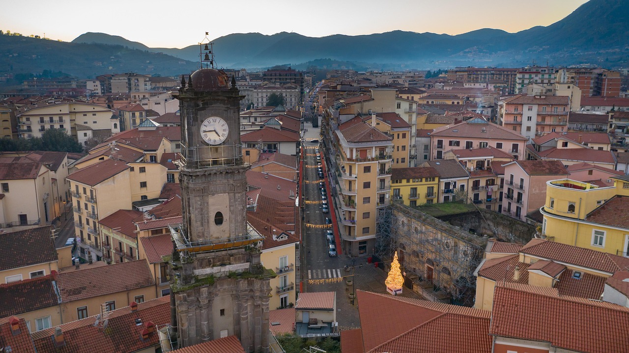 Aerial view of Avellino, Italy, featuring a historic clock tower, festive street lights, and surrounding rooftops with mountains in the background.